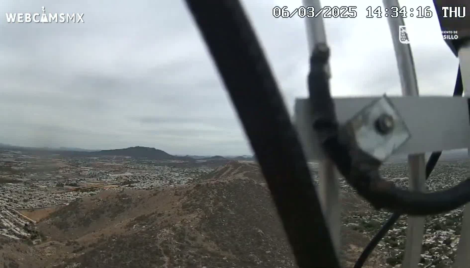 A view from a high vantage point shows a sprawling town below nestled in a hilly, desert-like landscape under an overcast sky.