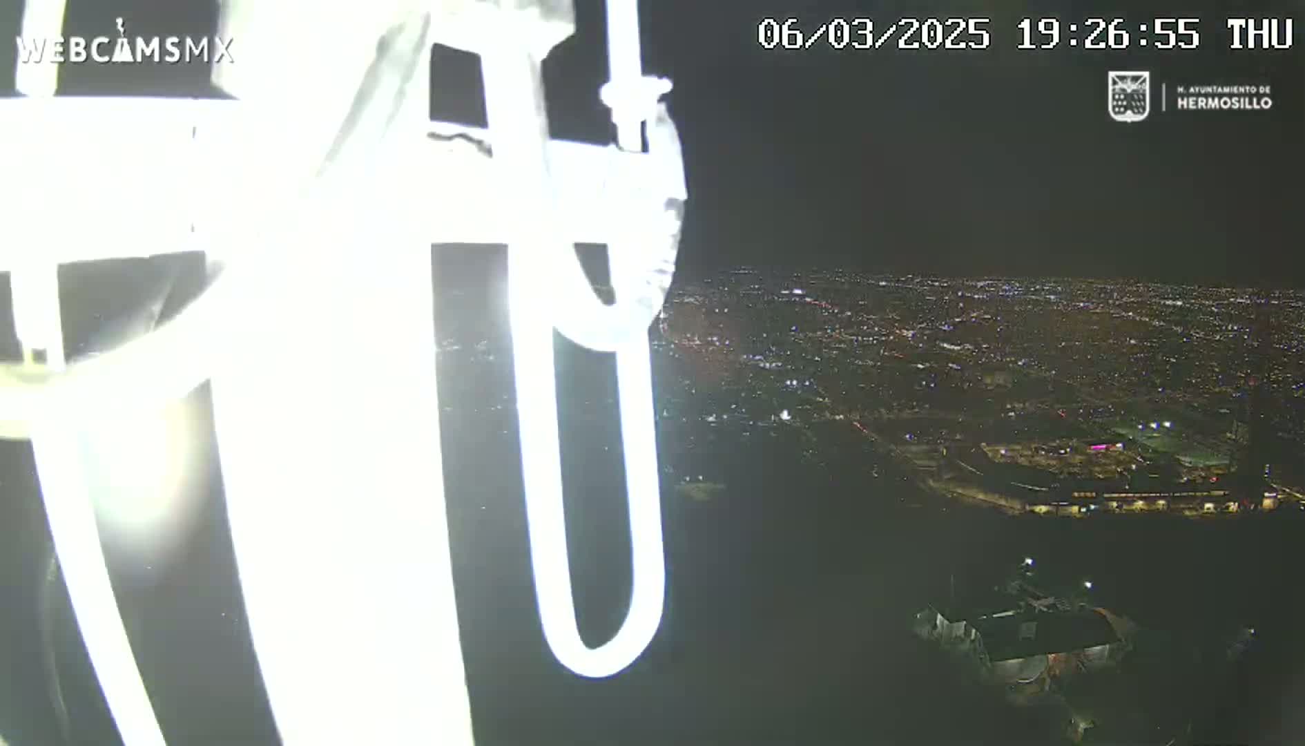A nighttime, high-angle view from a tall structure shows a brightly lit city sprawling in the distance.