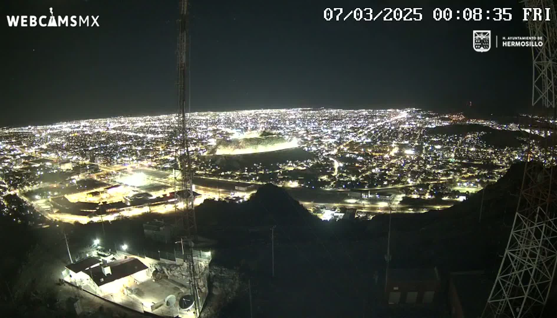 A nighttime, high-angle view of a sprawling city illuminated by lights, seen from a hilltop vantage point.