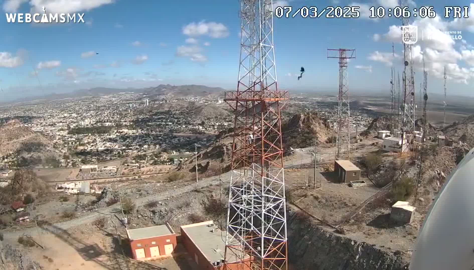 A high-angle view on a partly cloudy day shows several communication towers on a rocky hill overlooking a sprawling city.