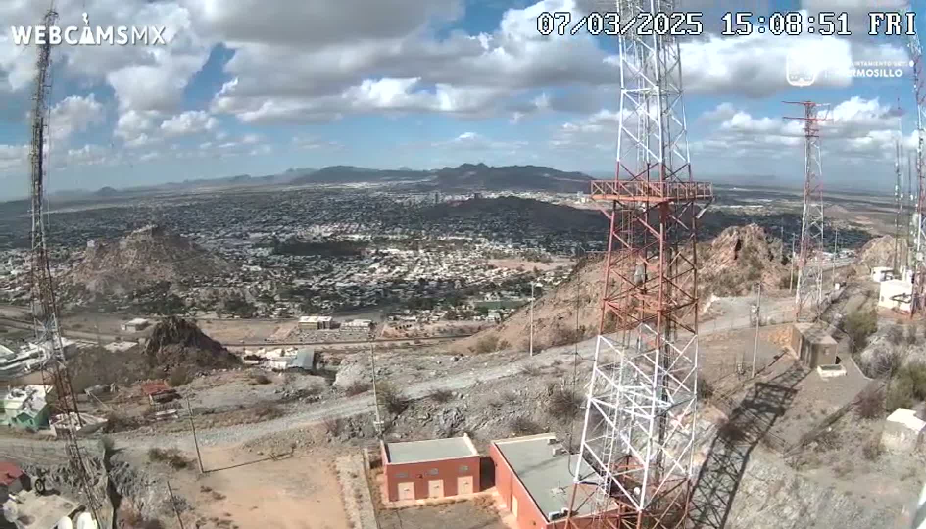 A high-angle view on a partly cloudy day shows a sprawling city below a hilltop with several communication towers, small buildings, and rocky terrain.