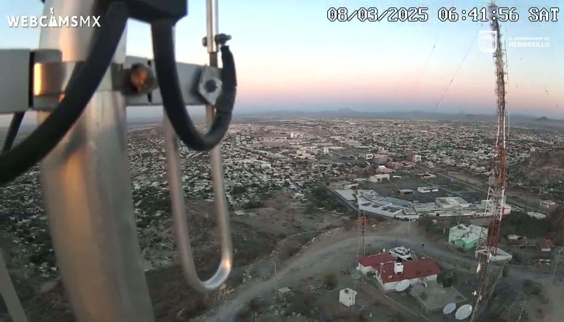 A high-angle, sunrise view of a city sprawling across a desert landscape, seen from atop a tower with communication equipment.