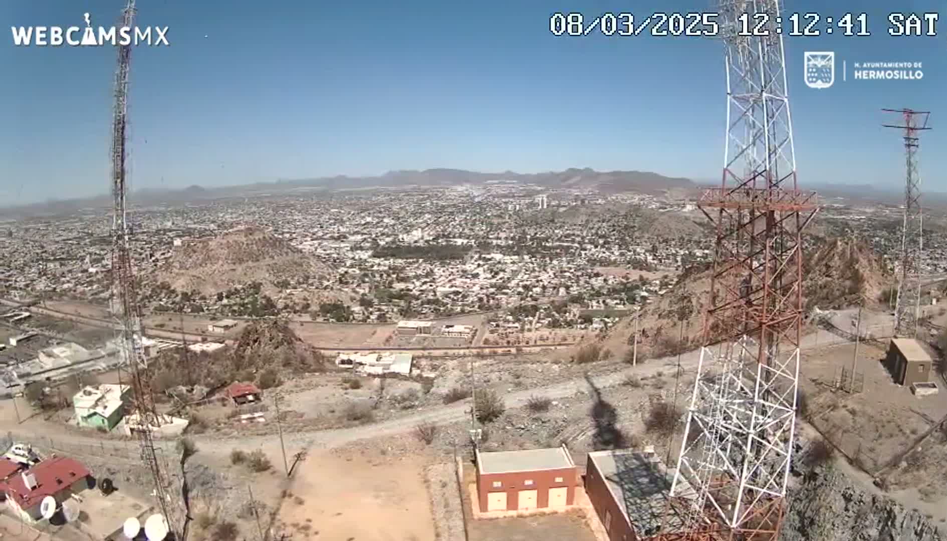 A high-angle, long shot on a clear, sunny day shows a large city sprawling across a valley below two tall communication towers perched atop a rocky hill.