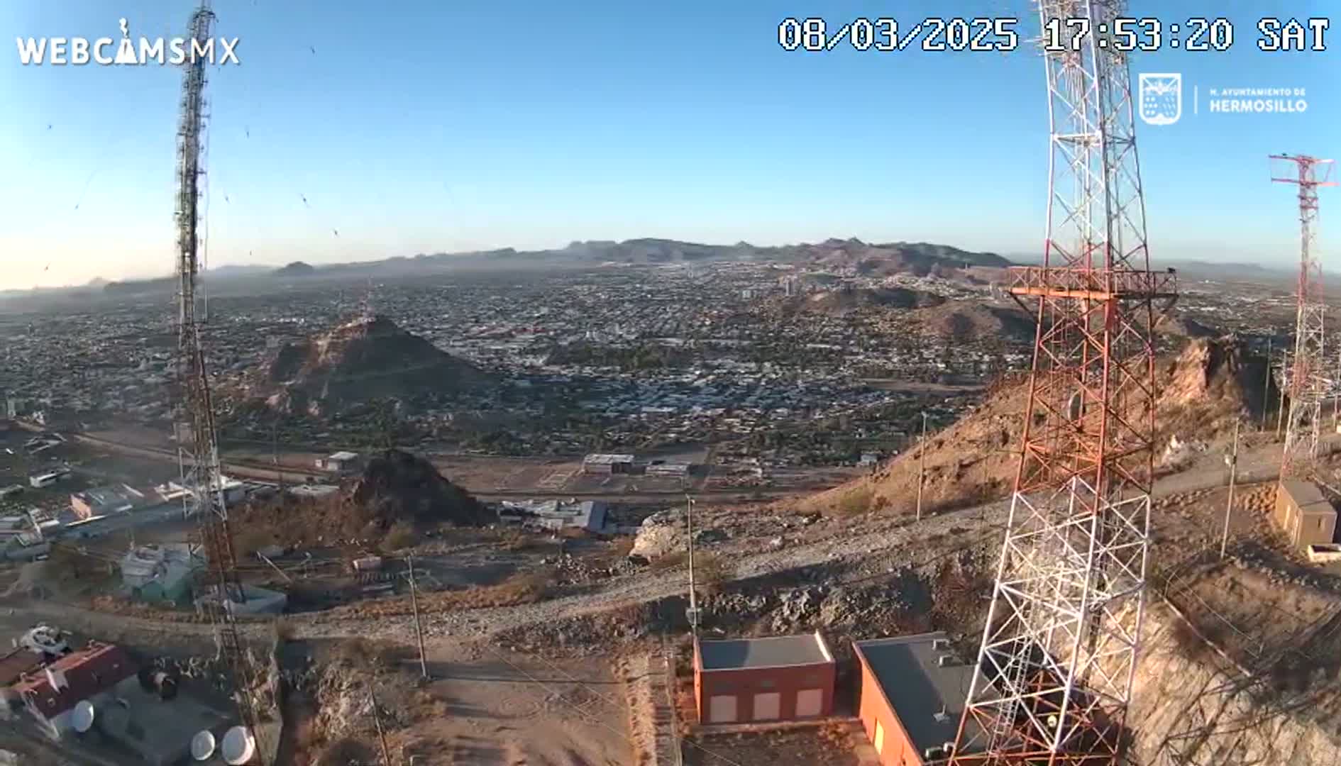 A high-angle, long shot on a clear day shows a sprawling city viewed from a hilltop, where two tall communication towers stand near some small buildings.