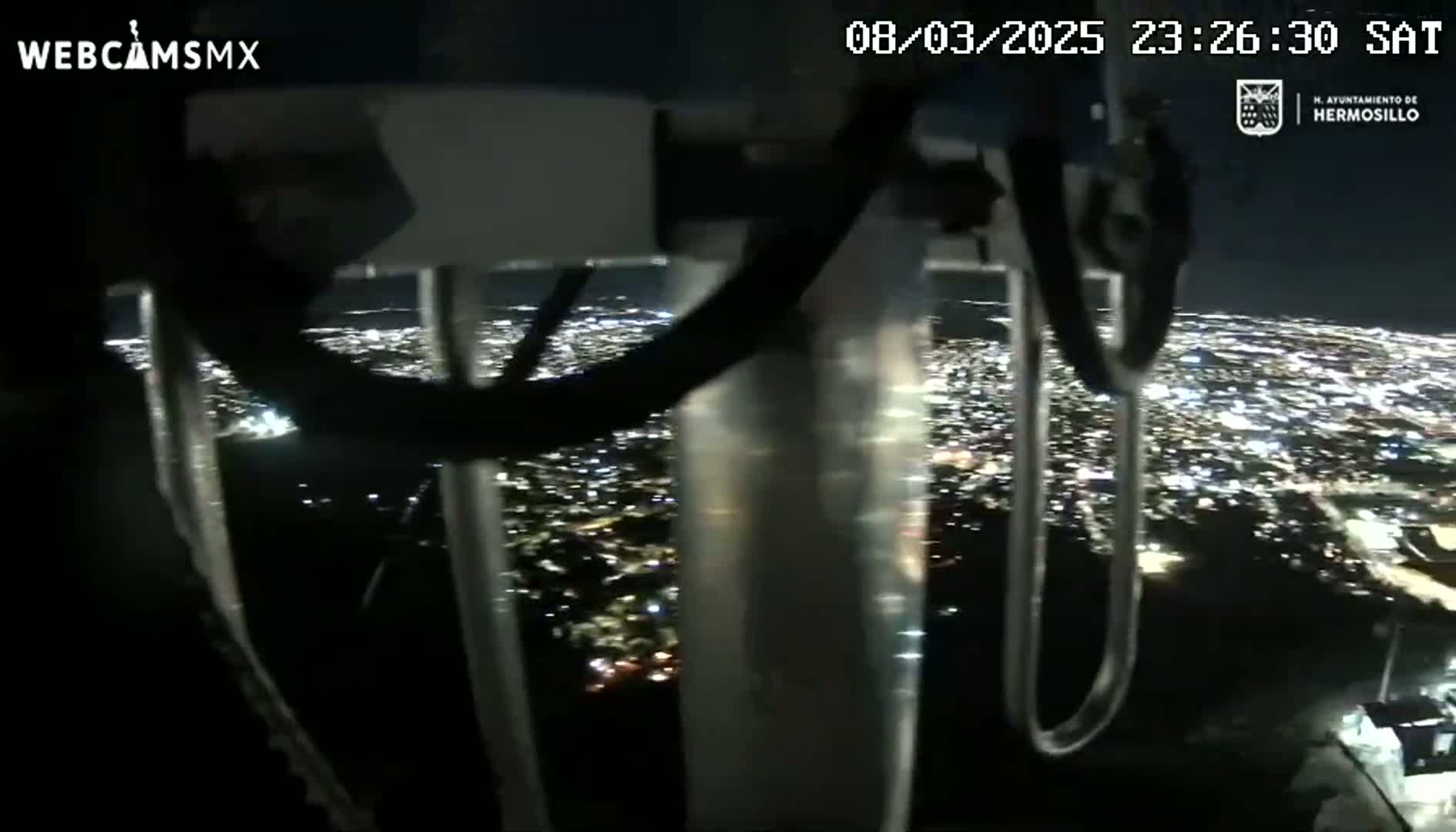 A nighttime view of a sprawling city illuminated by countless lights, seen from a high vantage point through the framework of what appears to be a tall structure.