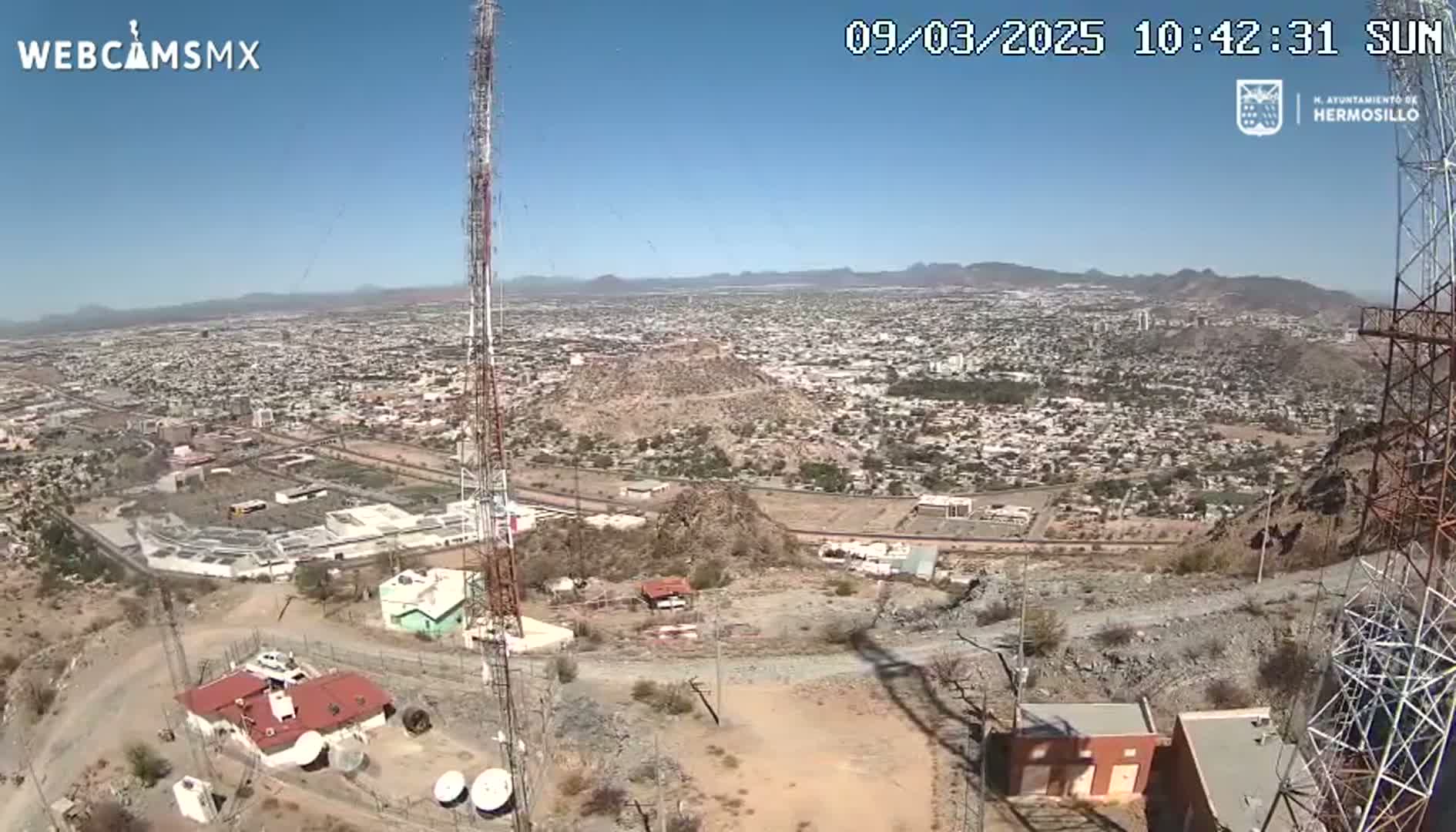 A high-angle, long shot on a sunny day shows a large city sprawling across a valley, viewed from a hilltop with communication towers and buildings.