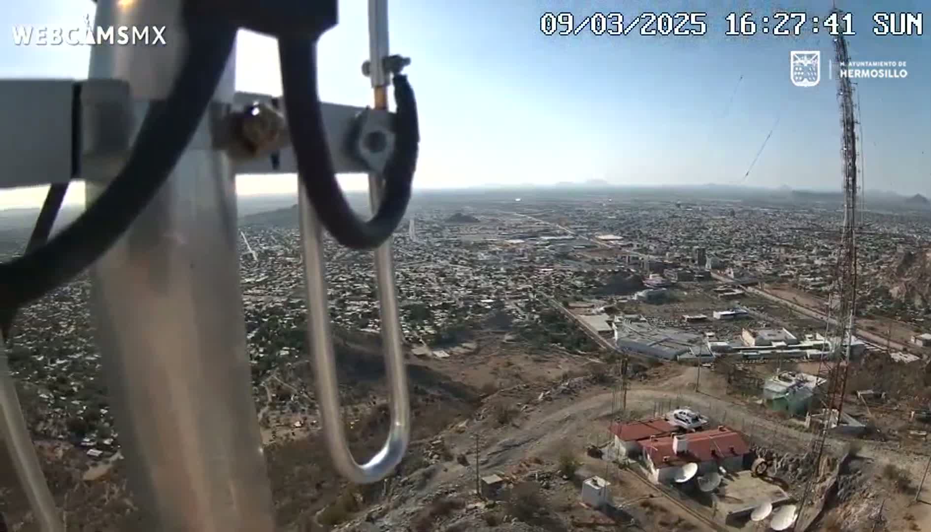 A high-angle, sunny view from a metal pole shows a sprawling city extending to distant mountains.