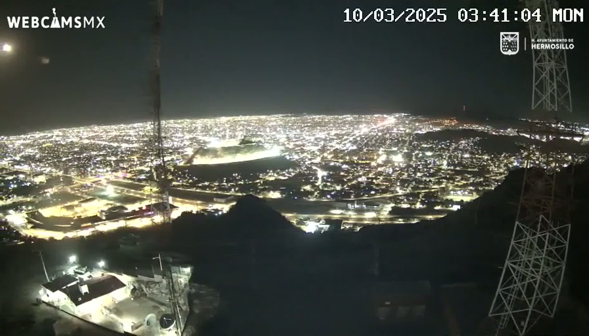 A nighttime, high-angle view shows a sprawling city illuminated below, seen from a vantage point on a hill or mountain.