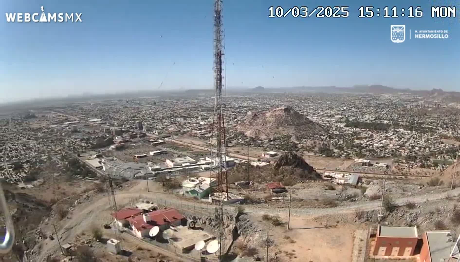 A high-angle view on a clear, sunny day shows a sprawling city below a hilltop with communication towers and buildings, along with rocky, arid terrain.