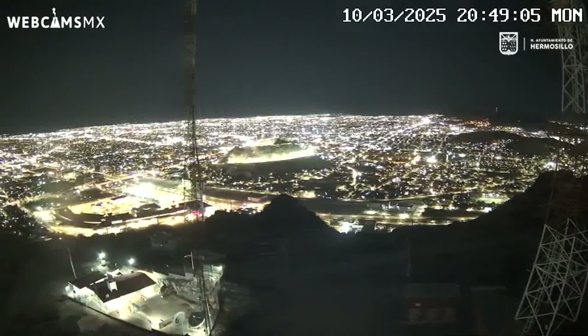 A nighttime, high-angle view shows a sprawling city illuminated by countless lights, with a portion of a dark mountainside in the foreground.