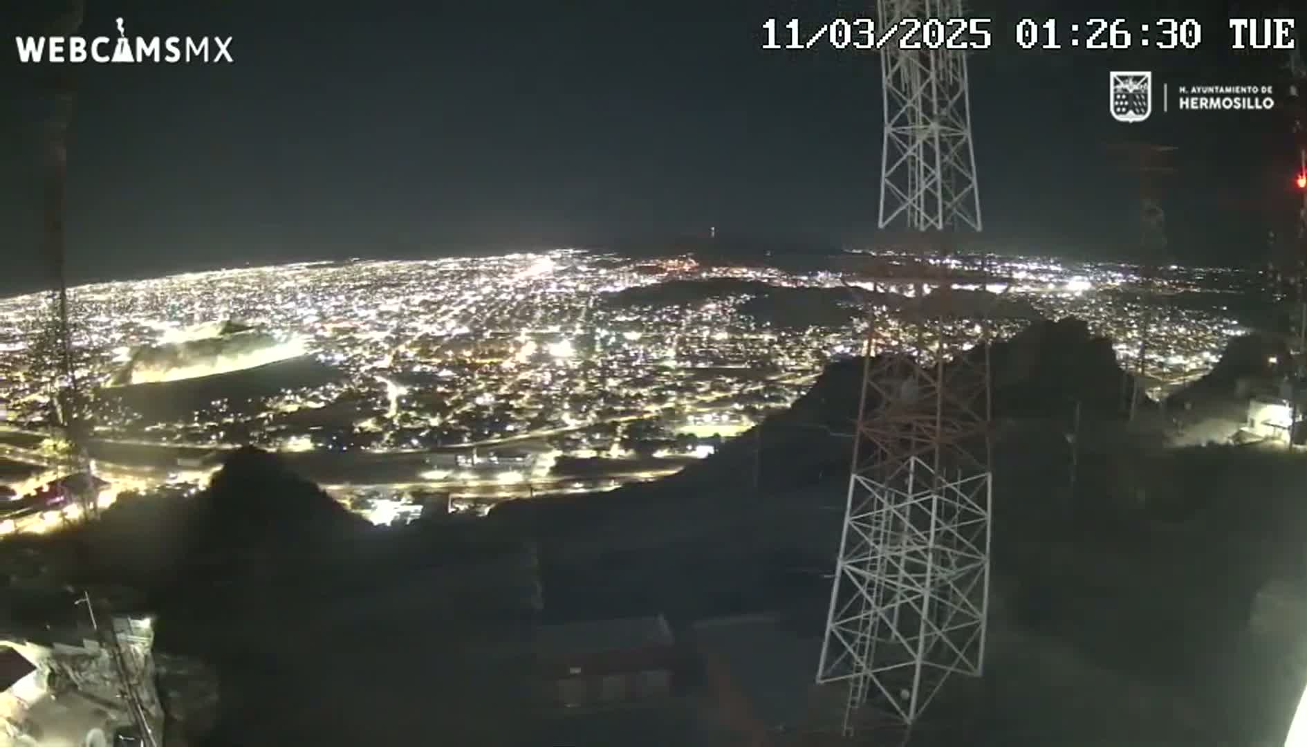 A nighttime, high-angle view shows a sprawling city illuminated by lights, observed from a hilltop near communication towers under a clear sky.