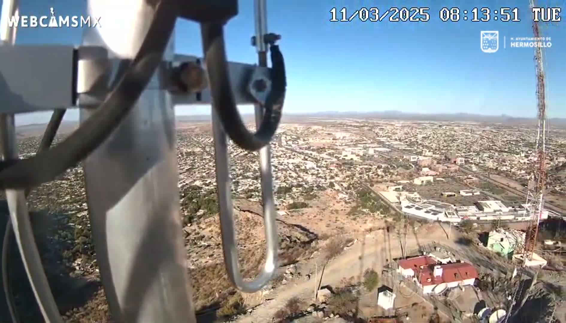A high-angle view on a clear, sunny day shows a sprawling city below, seen from a vantage point near a tall antenna tower.