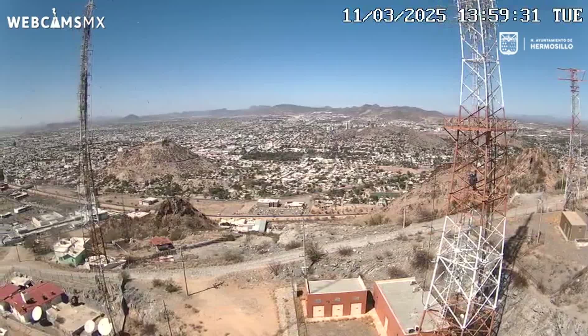 A high-angle, sunny view shows a sprawling city extending to distant mountains, with two tall communication towers standing prominently on a rocky hillside.
