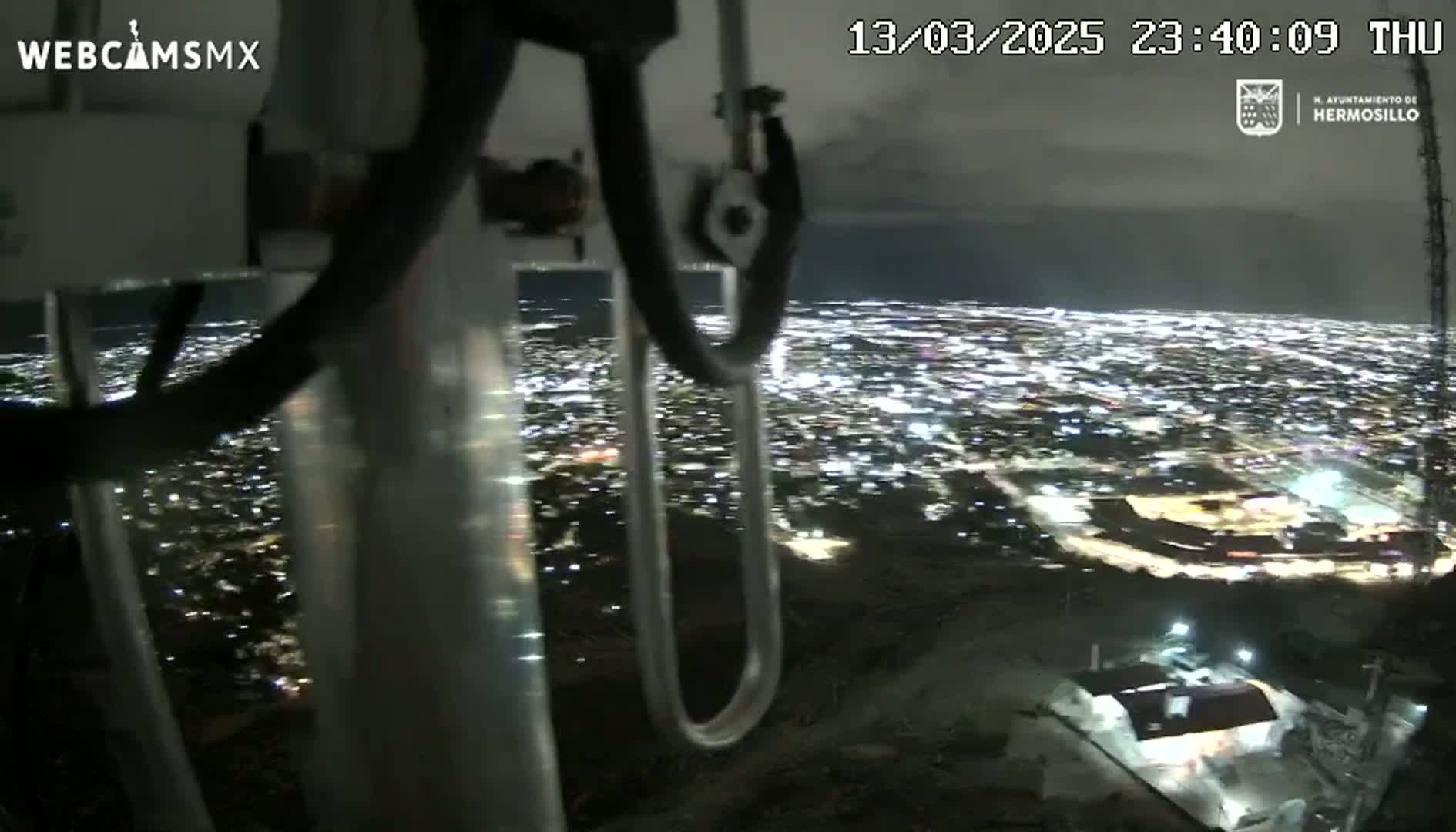 A nighttime, slightly cloudy view of a city sprawling below, seen from a high vantage point through the metal framework of what appears to be a camera mount.