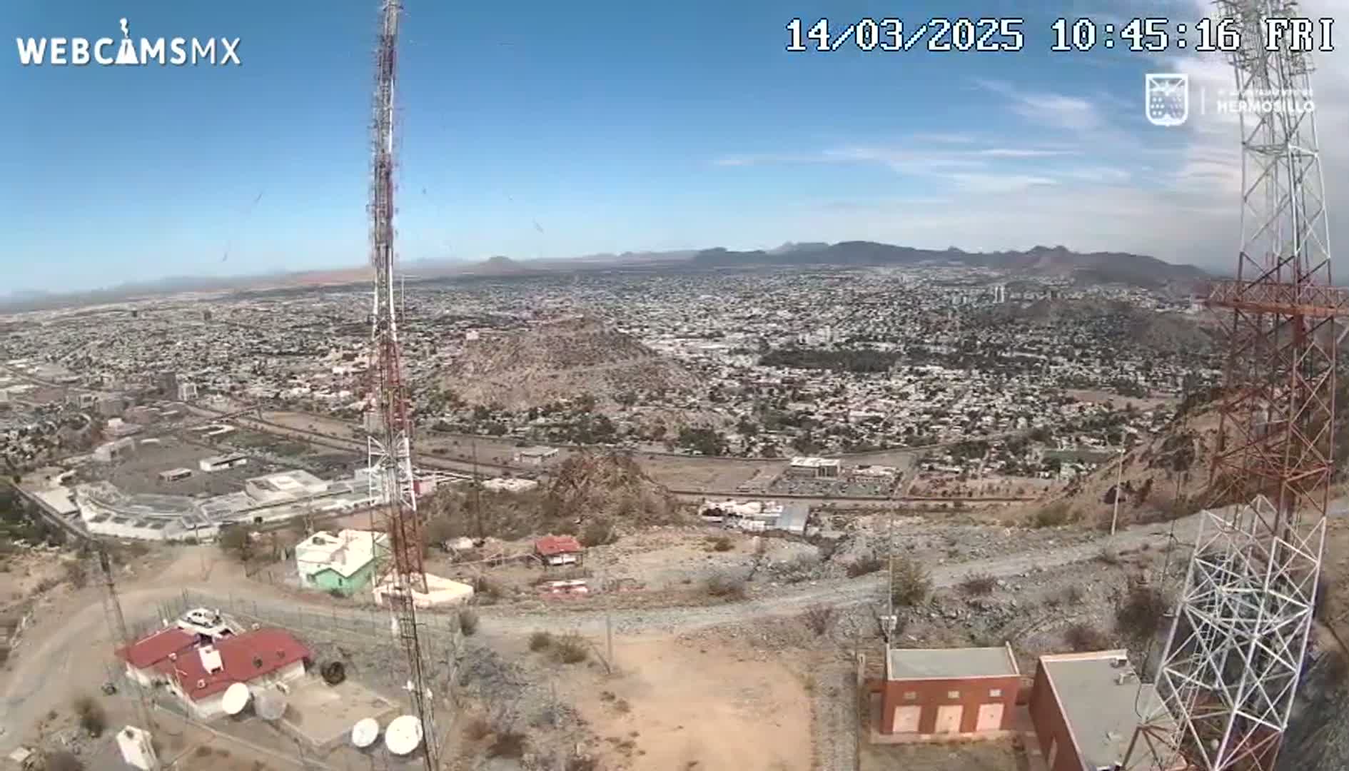 A high-angle, long shot on a clear, sunny day shows a sprawling city below a rocky hilltop where two communication towers stand, along with small buildings and satellite dishes.