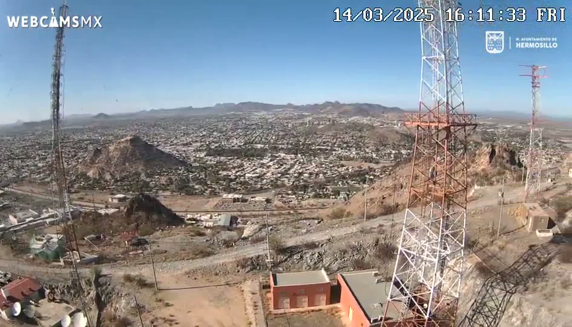 A high-angle, sunny view shows a sprawling city below, hillsides with sparse vegetation, and several tall communication towers on a rocky, dusty hillside.