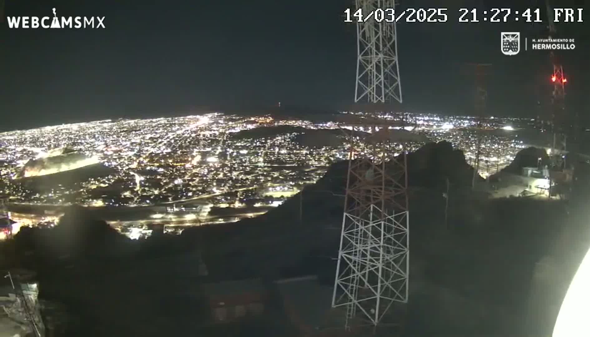 A nighttime view from a hilltop shows a brightly lit city sprawling below, with two tall communication towers in the foreground under a clear sky.