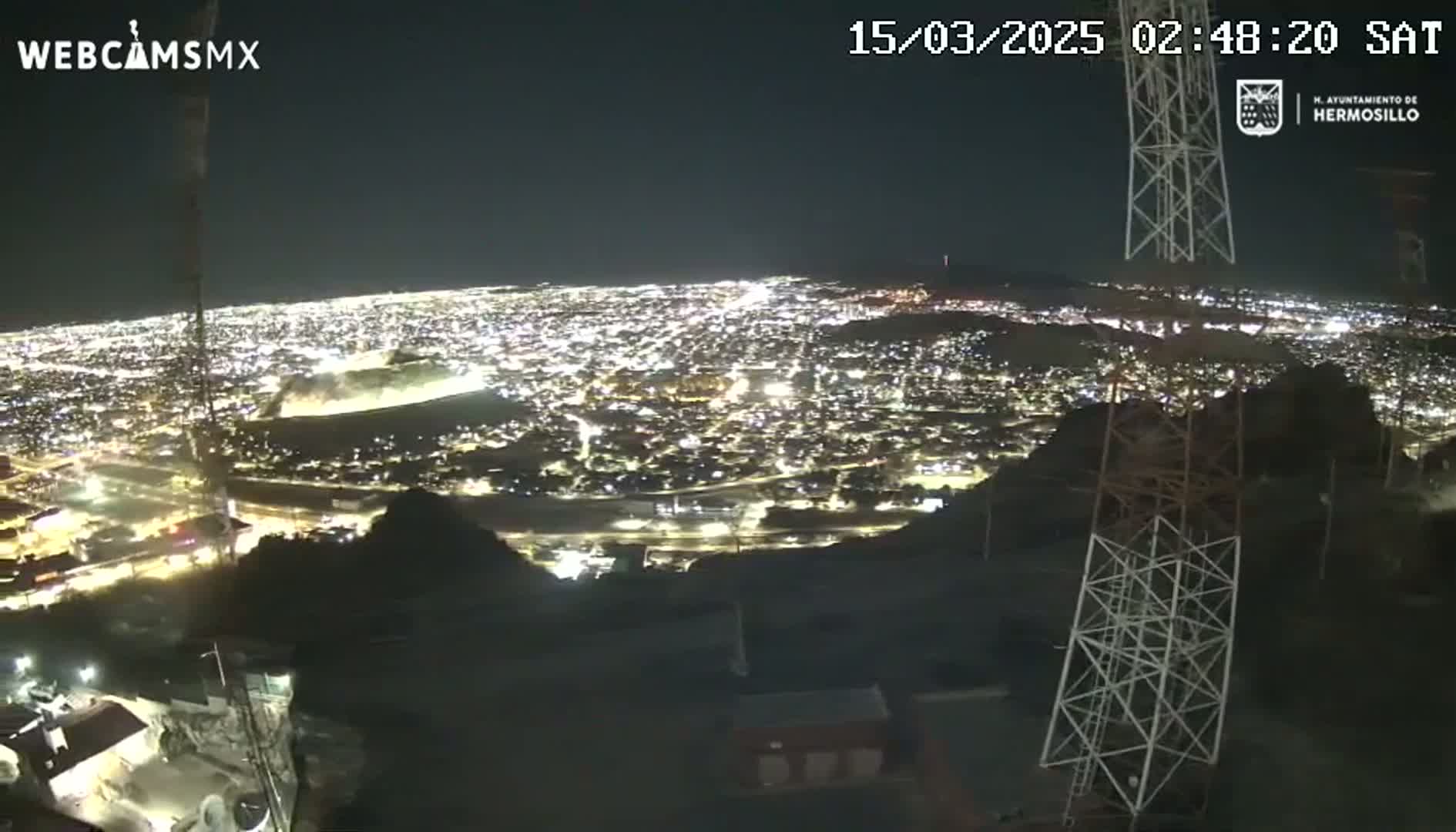 A nighttime, high-angle view shows a sprawling city illuminated below, with several tall communication towers in the foreground.