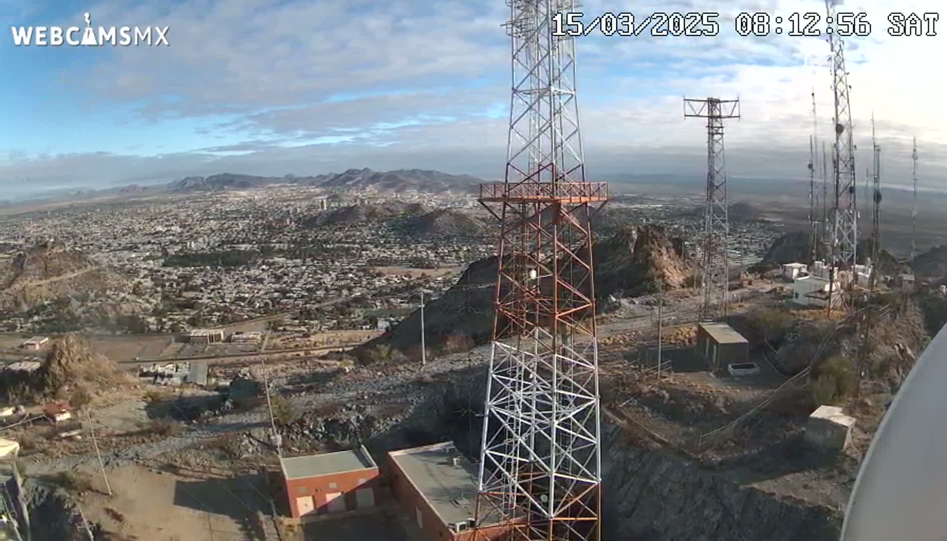 A high-angle, long shot on a clear, sunny day shows several communication towers atop a rocky hill overlooking a sprawling city nestled in a mountainous region.