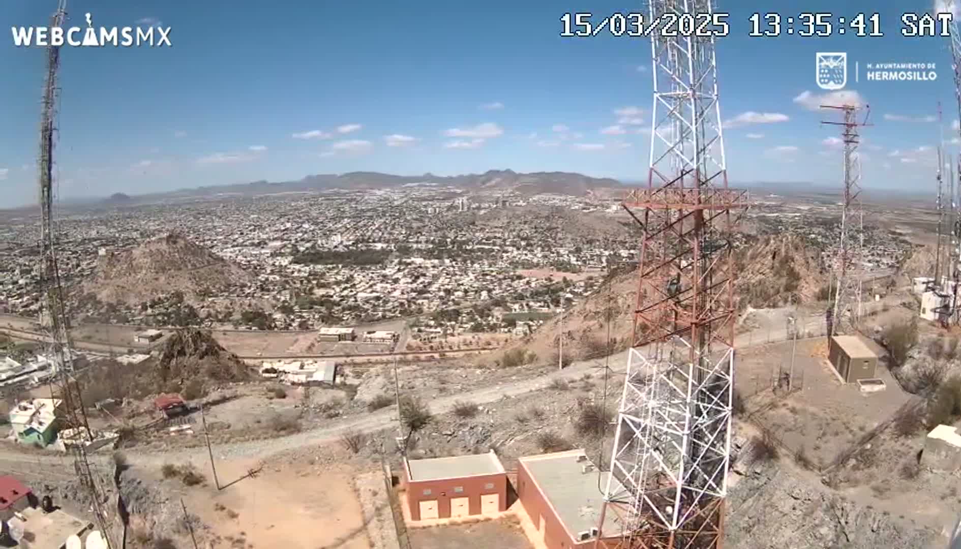 A high-angle, sunny view from a hilltop shows a sprawling city below, with communication towers and small buildings nearby.