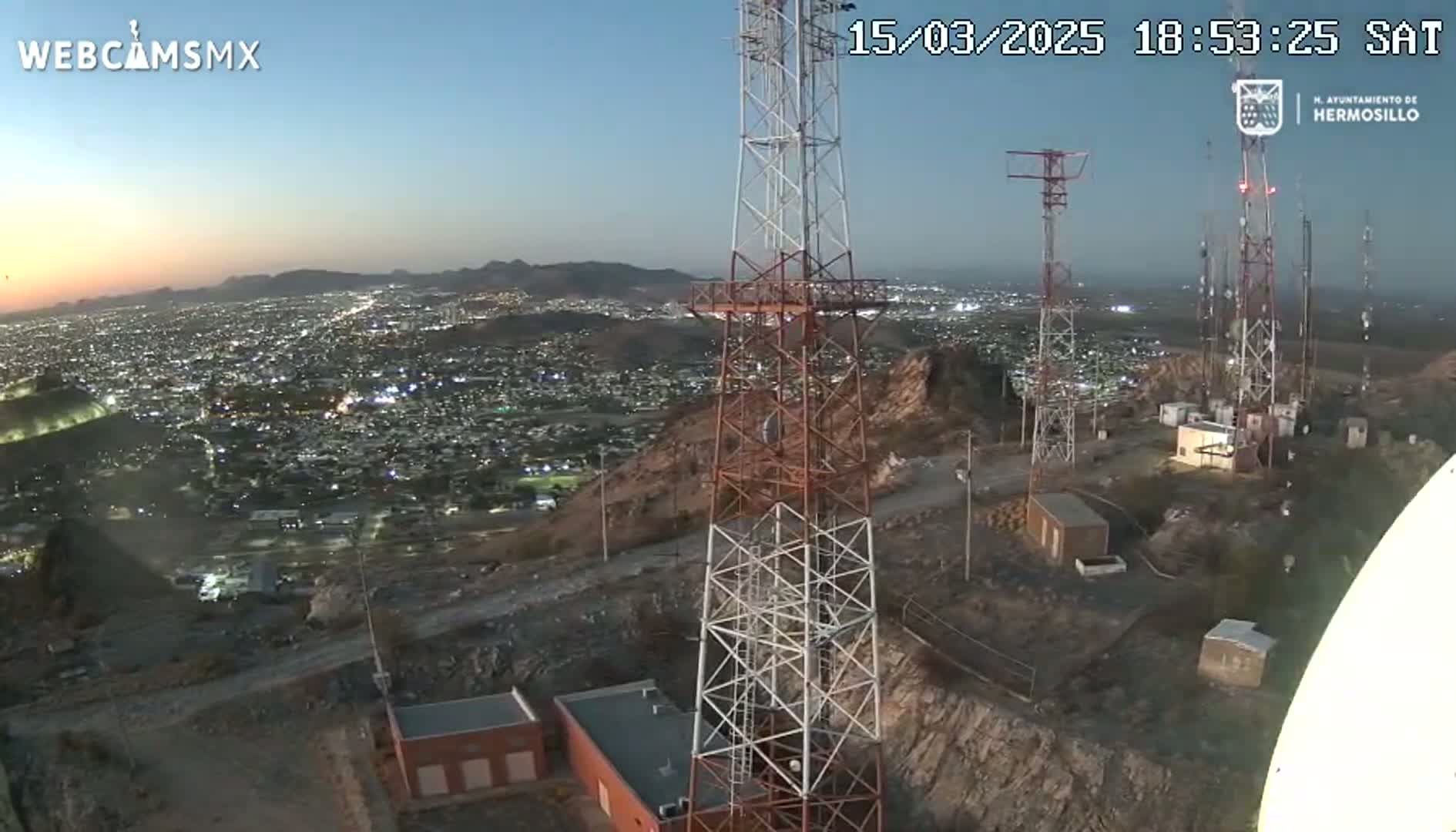 A nighttime view from a hilltop shows several communication towers overlooking a sprawling city illuminated by lights under a twilight sky.