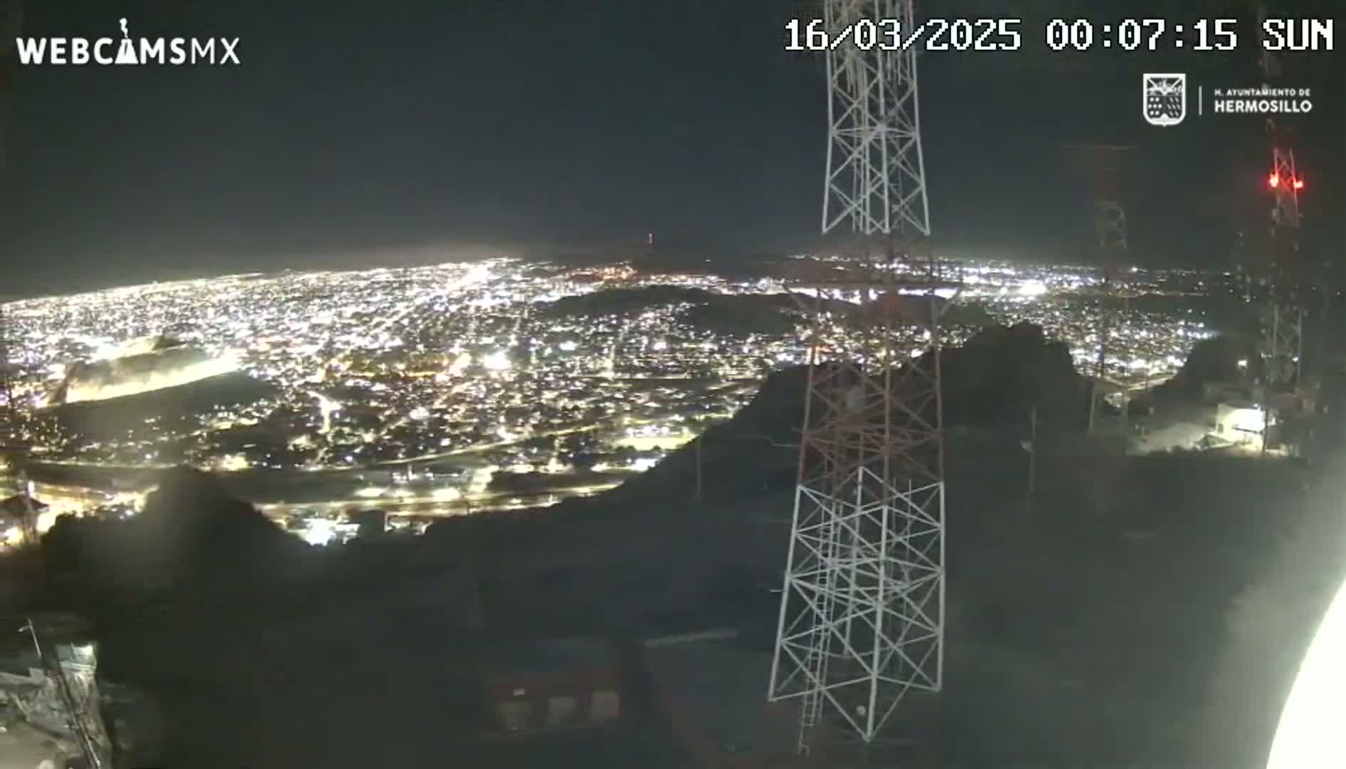 A nighttime view from a hilltop overlooking a brightly lit city, with several tall communication towers in the foreground under a clear sky.