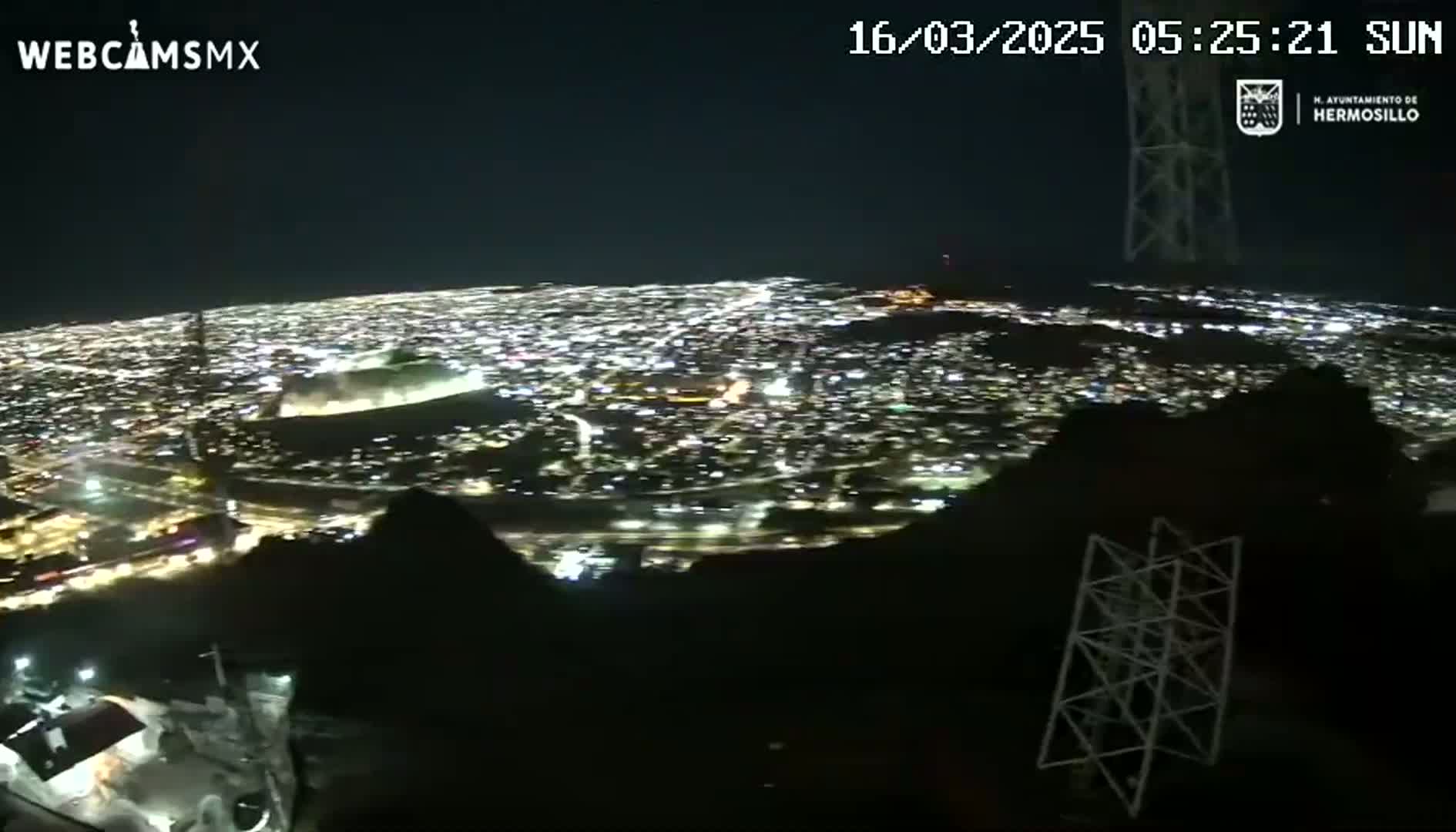 A nighttime, high-angle view shows a sprawling city illuminated below, partially obscured by dark hills and structures in the foreground.
