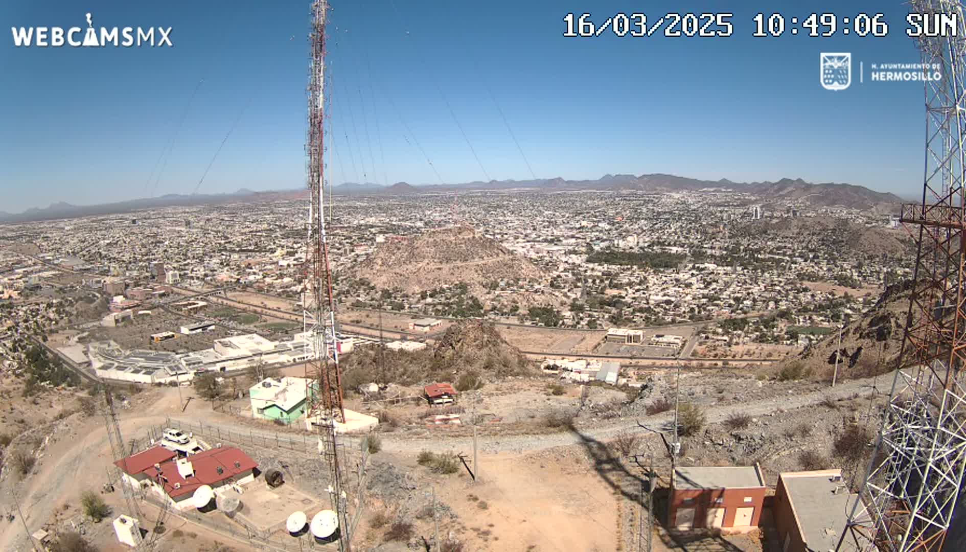 A high-angle, sunny view of a sprawling city nestled in a mountainous region, with communication towers and buildings situated on a rocky hillside overlooking the urban landscape.