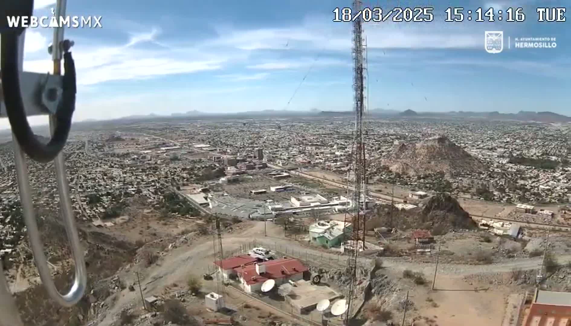 A high-angle, long shot on a clear, sunny day shows a sprawling city below a hilltop with communication towers and small buildings.
