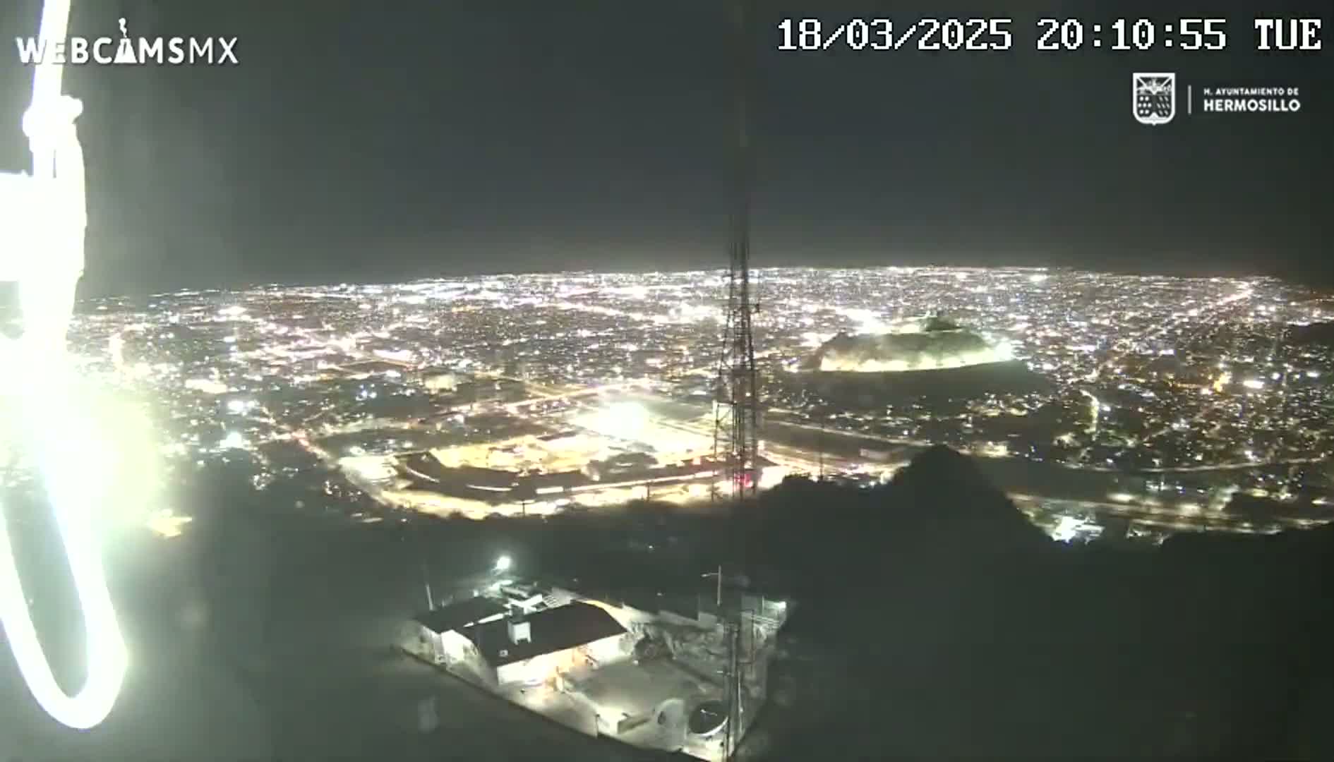 A nighttime, high-angle view shows a sprawling city illuminated by numerous lights, with a tall communication tower in the foreground and a dark hill or mountain in the middle ground.