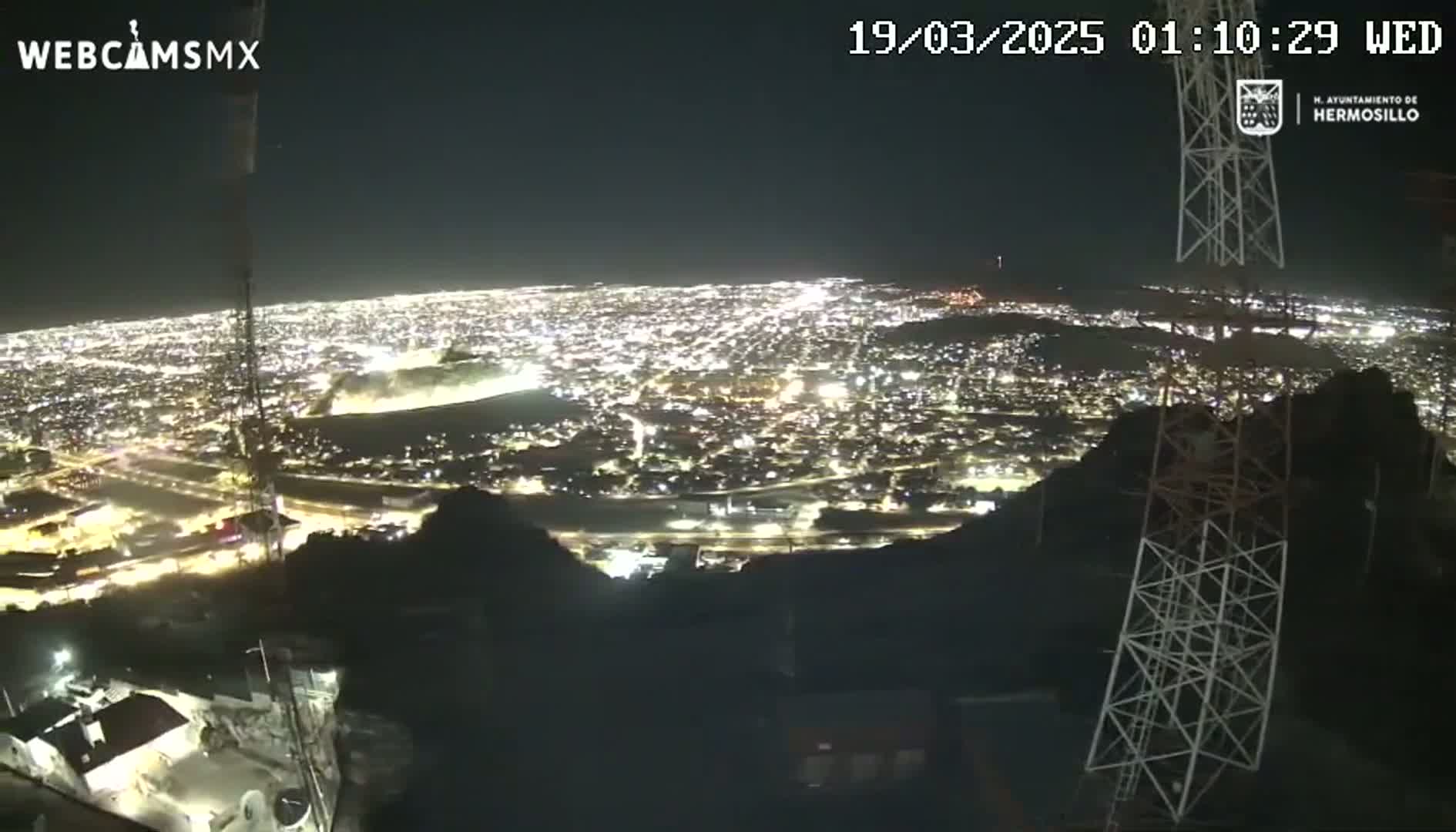 A nighttime view from a hilltop shows a sprawling city illuminated below, under a clear sky.