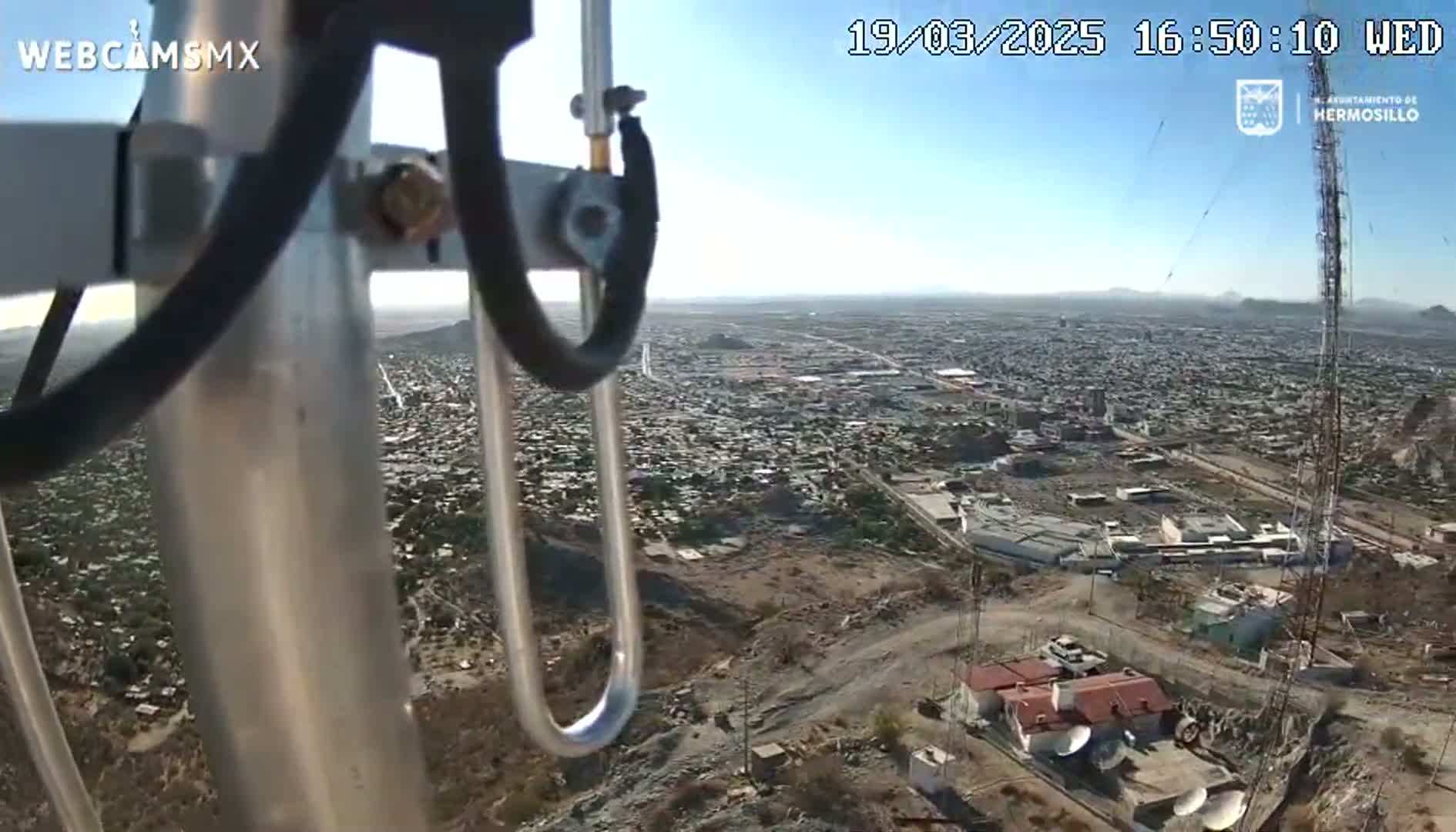 A high-angle, outdoor view on a clear day shows a sprawling city below, partially obscured by a metal pole and wires in the foreground.