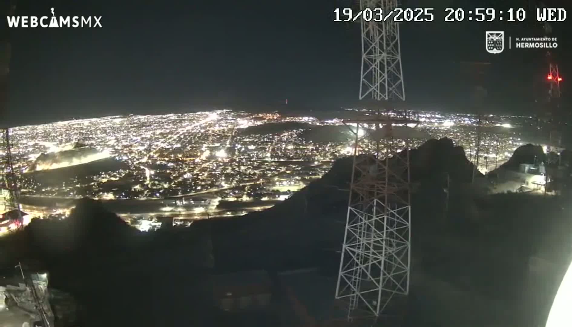 A nighttime, aerial view of a sprawling city illuminated by countless lights, seen from a vantage point on a hilltop with communication towers in the foreground.