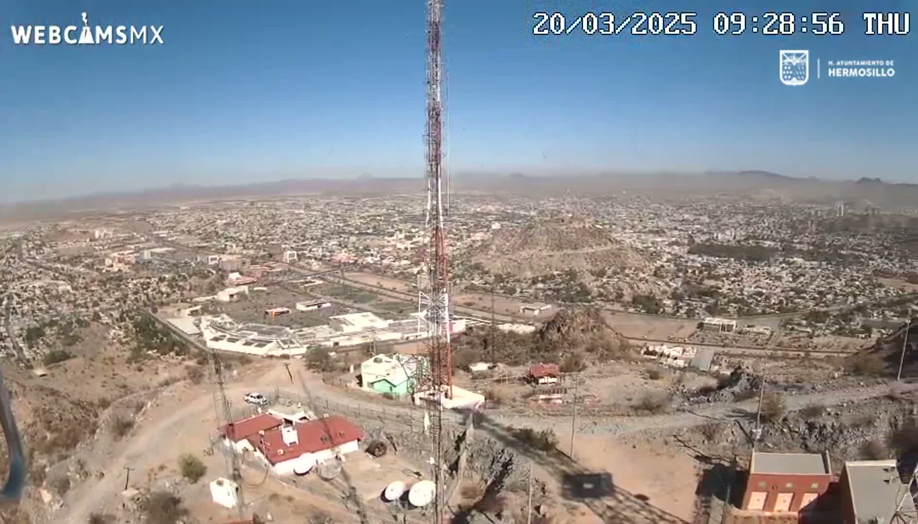 A tall communication tower stands atop a hill overlooking a sprawling city under a clear, sunny sky.