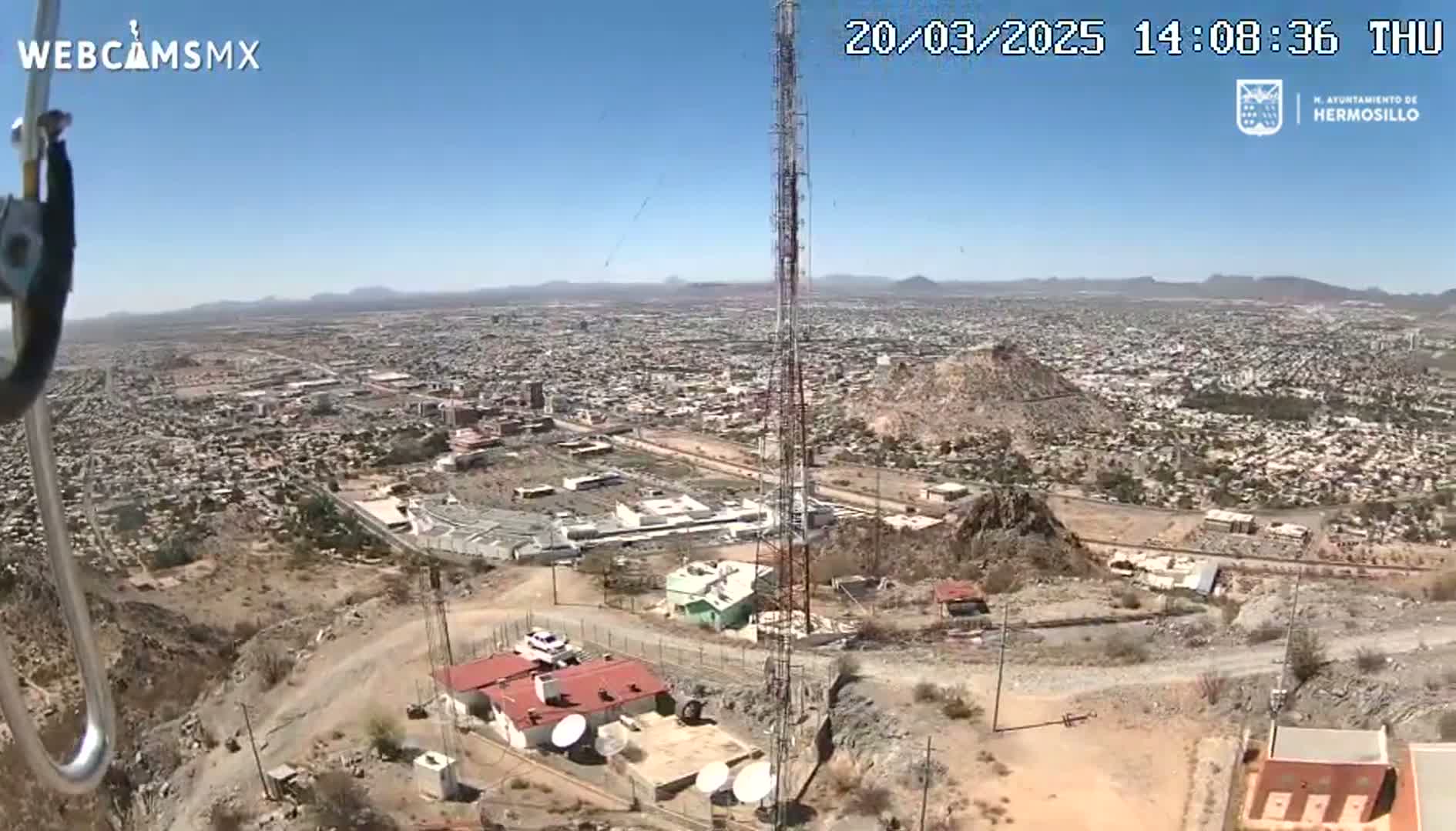 A high-angle, long shot on a clear, sunny day shows a sprawling city below a rocky hilltop with a communications tower and several buildings.