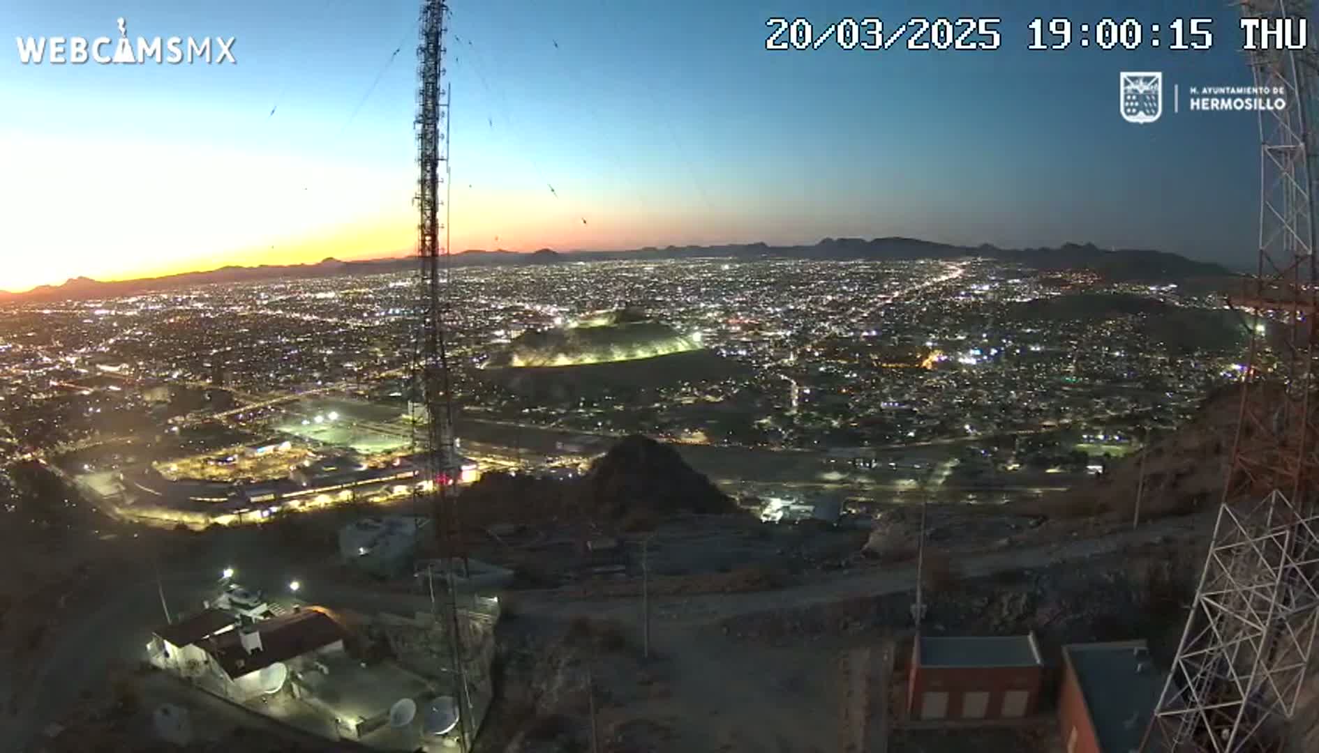 A nighttime, twilight view from a hilltop overlooking a sprawling city illuminated by lights, with a tall communication tower prominently featured in the foreground.