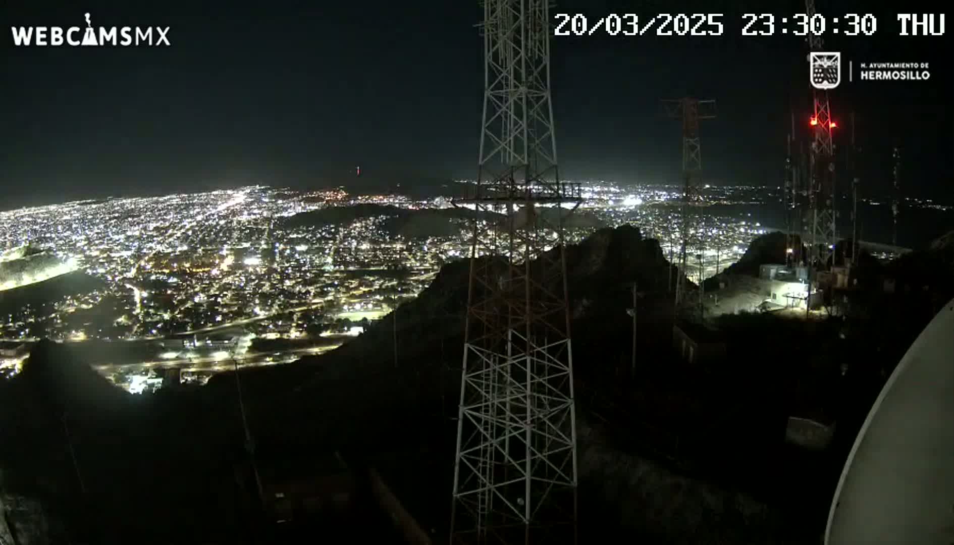 A nighttime, high-angle view shows a sprawling city illuminated below, with several tall communication towers silhouetted against the dark sky in the foreground.
