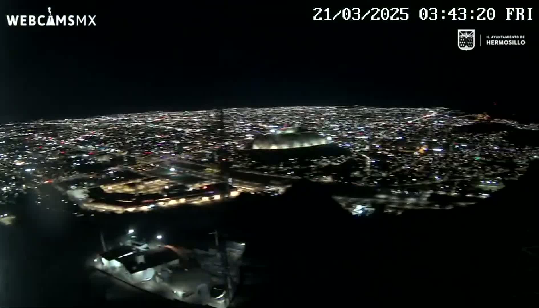 A nighttime, high-angle view shows a sprawling city illuminated by countless lights, with a large stadium visible in the mid-ground.