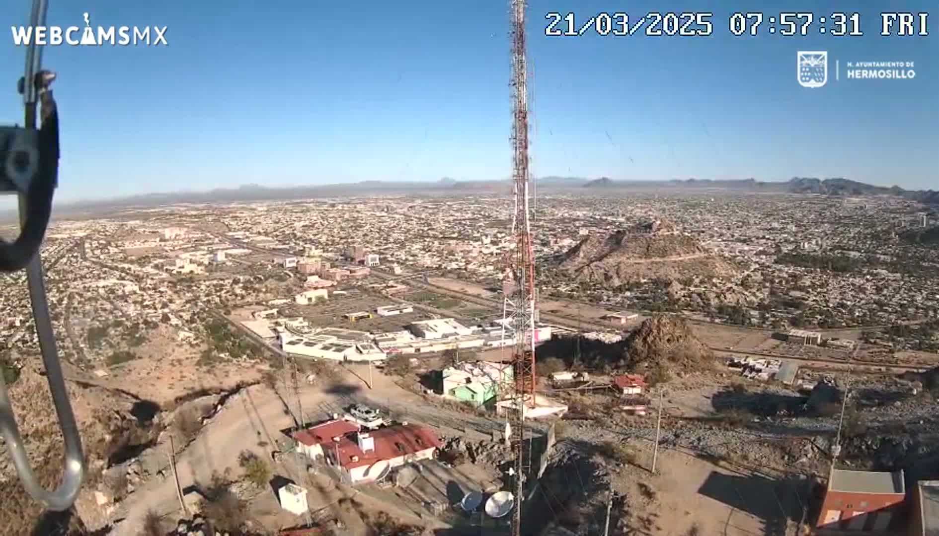 A high-angle, long shot on a sunny day shows a sprawling city below a rocky hillside with a tall communication tower and some buildings nearby.