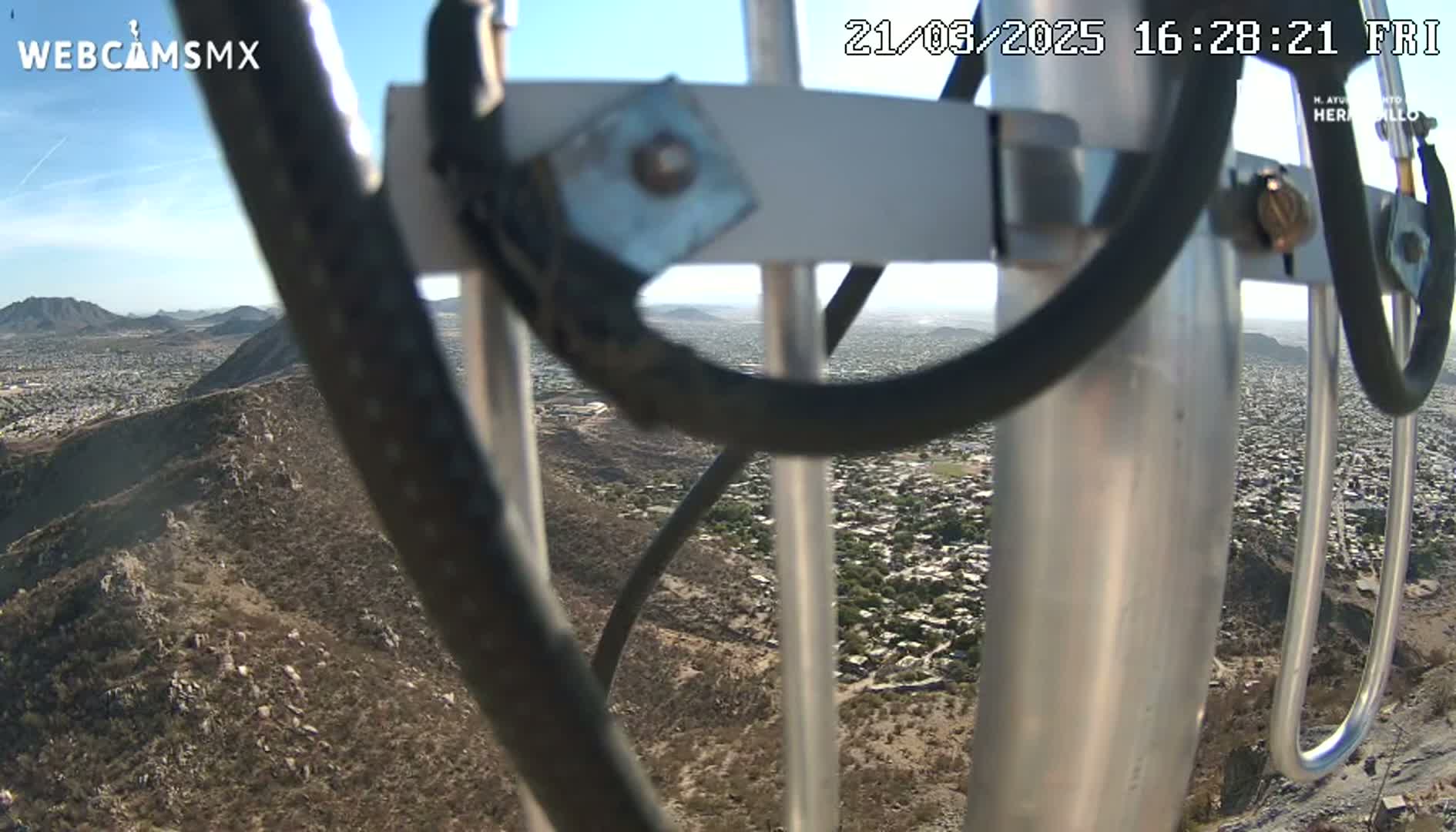A view of a city and surrounding hills from a high vantage point on a clear, sunny day, partially obscured by a metal antenna structure.