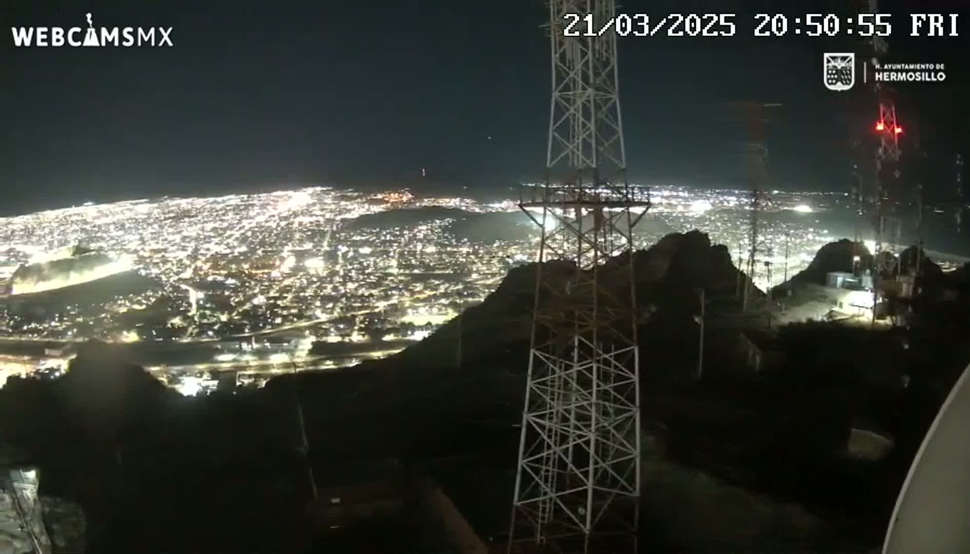 A nighttime view from a hilltop shows a brightly lit city sprawling below, with several tall communication towers nearby under a clear, dark sky.