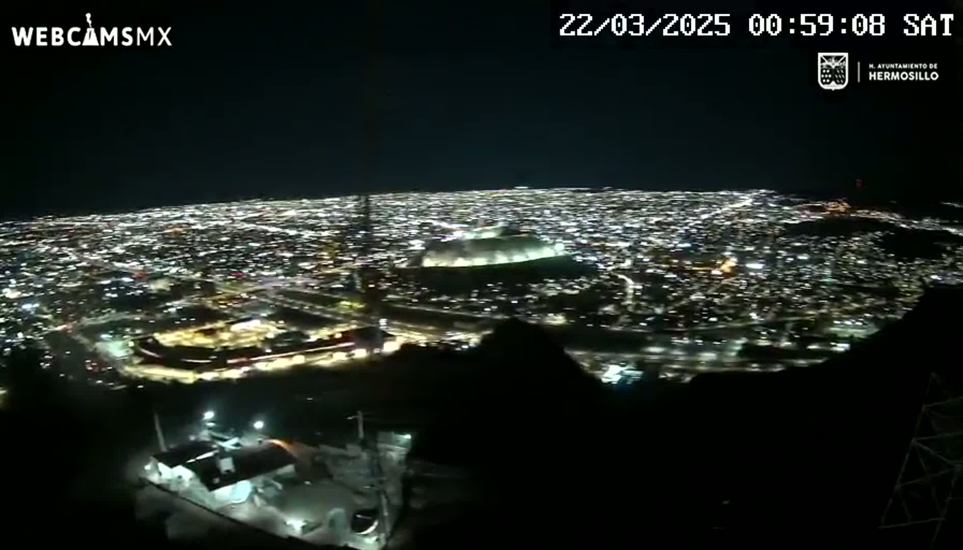 A nighttime, high-angle view shows a sprawling city illuminated by countless lights, including a large stadium, from a vantage point on a hill or mountain.