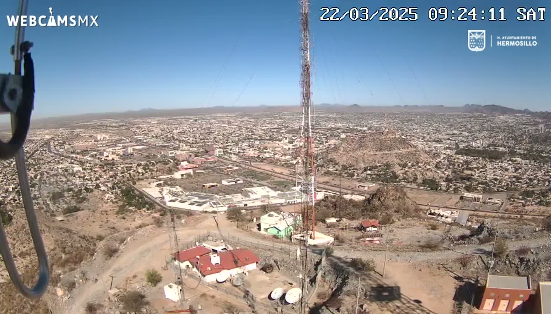 A high-angle, long shot on a clear, sunny day shows a large city sprawling across a valley, with a communication tower and buildings situated on a rocky hillside overlooking it.