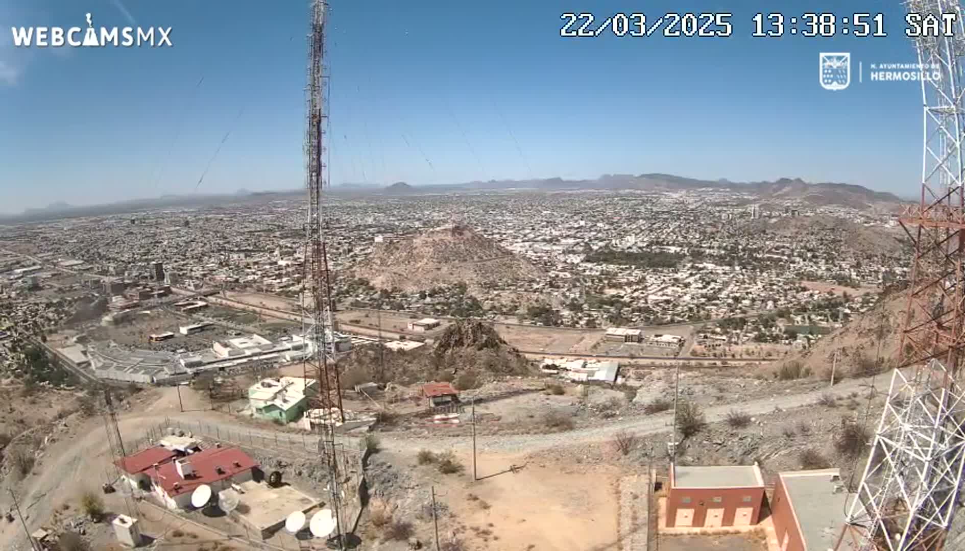 A high-angle, long shot on a clear, sunny day shows a sprawling city situated in a mountainous, desert-like region, with communication towers in the foreground.