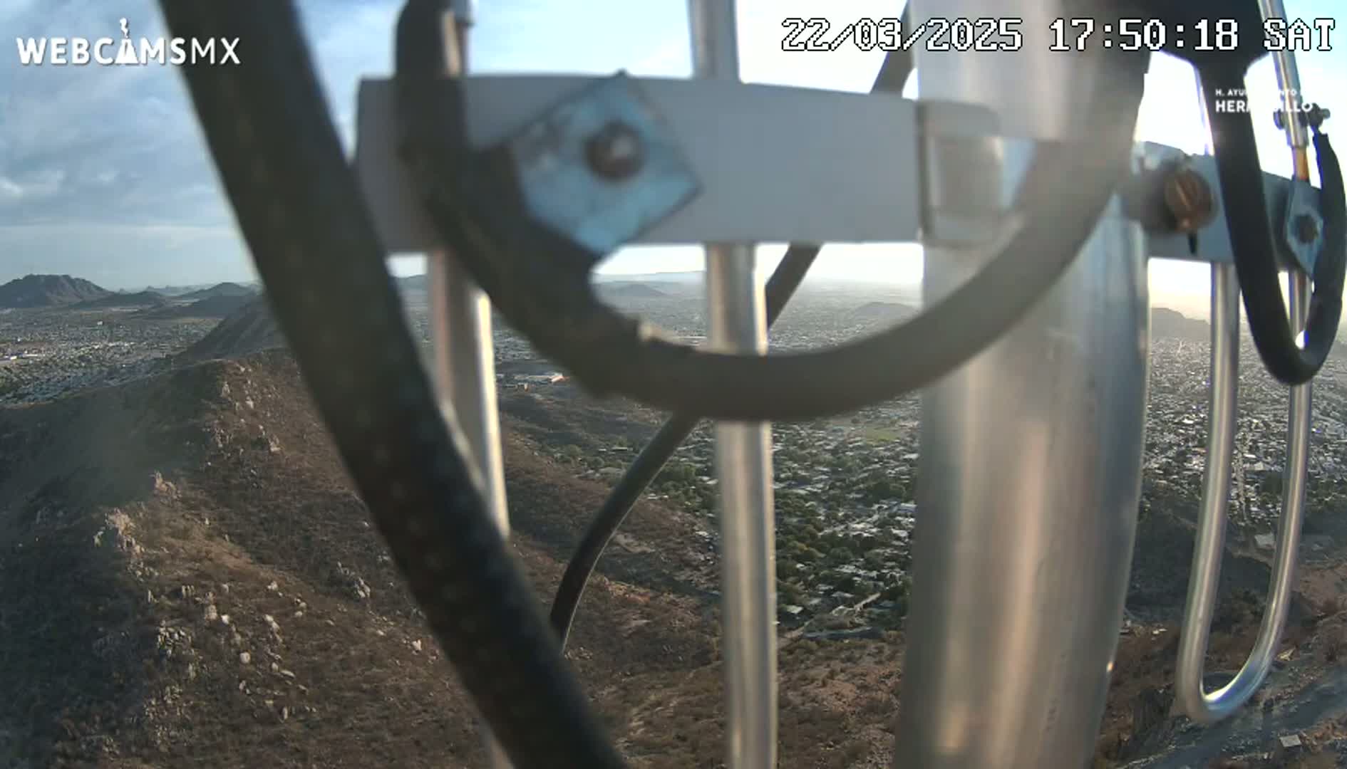 A view of a city and surrounding hills from a high vantage point on a partly cloudy day, seen through the metal framework of a camera mount.