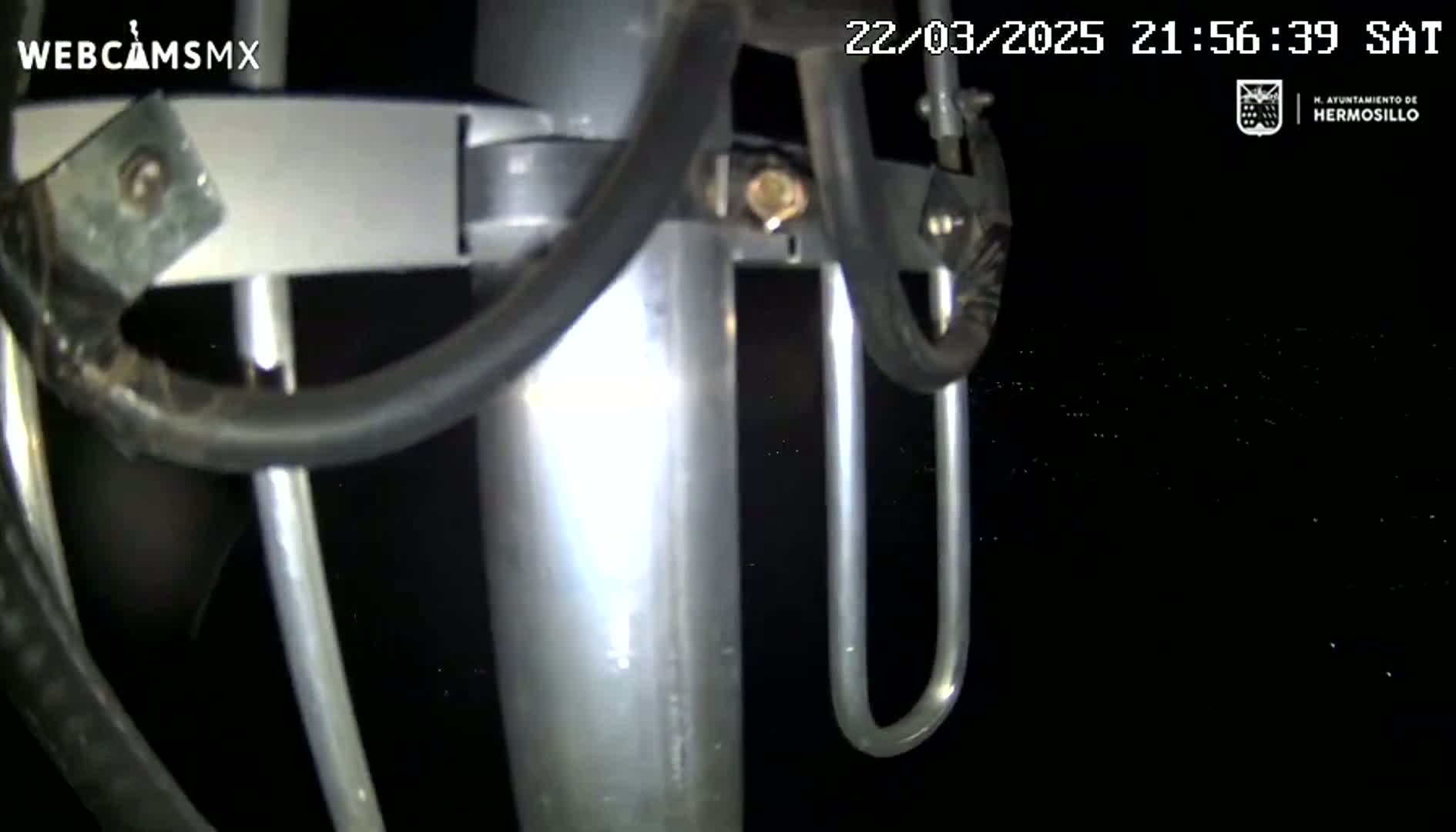 A close-up view at night shows a metallic pole with attached wires and brackets against a dark background speckled with faint lights.