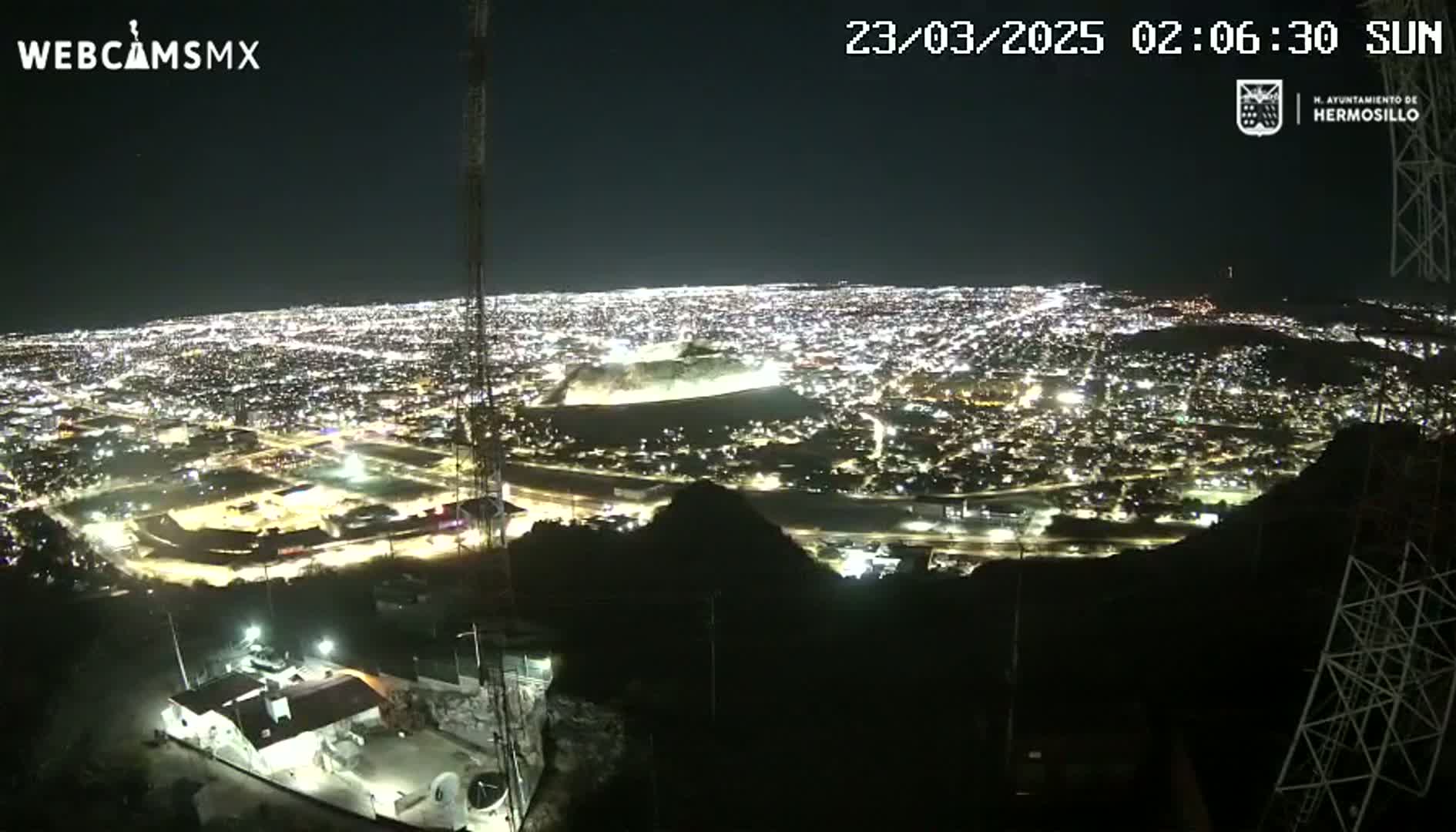 A nighttime view from a hilltop shows a sprawling city illuminated by many lights under a clear sky.