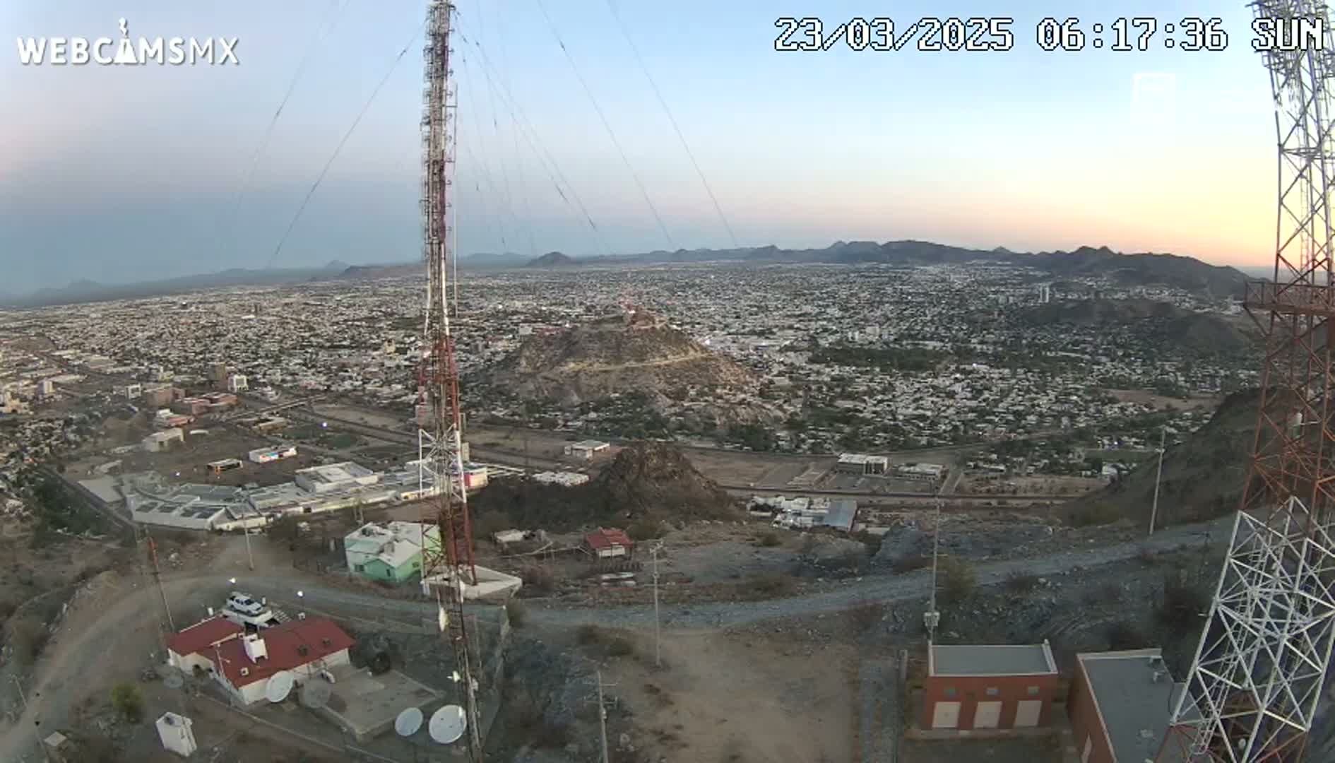 An expansive view of a city sprawls beneath a clear, sunrise sky, seen from a hilltop with communication towers and small buildings in the foreground.