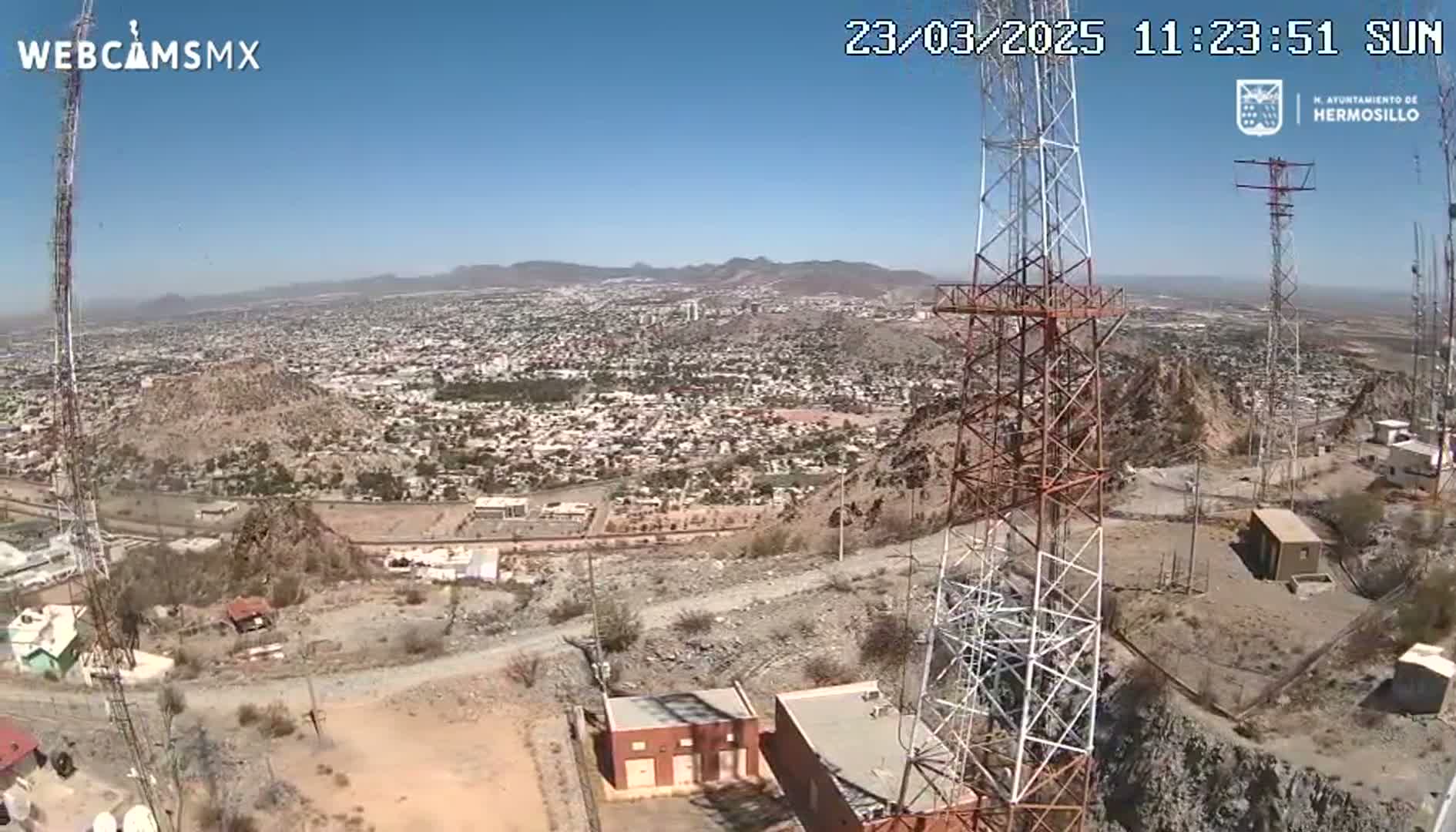 A sunny, high-angle view shows a sprawling city below several tall communication towers situated on a rocky, arid hillside.
