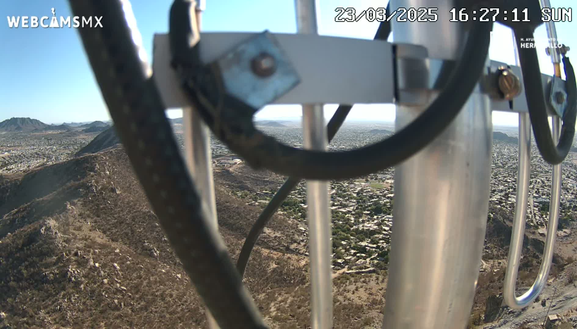 A view of a sprawling city and surrounding desert hills is seen through the metal framework of a tall antenna on a sunny day.
